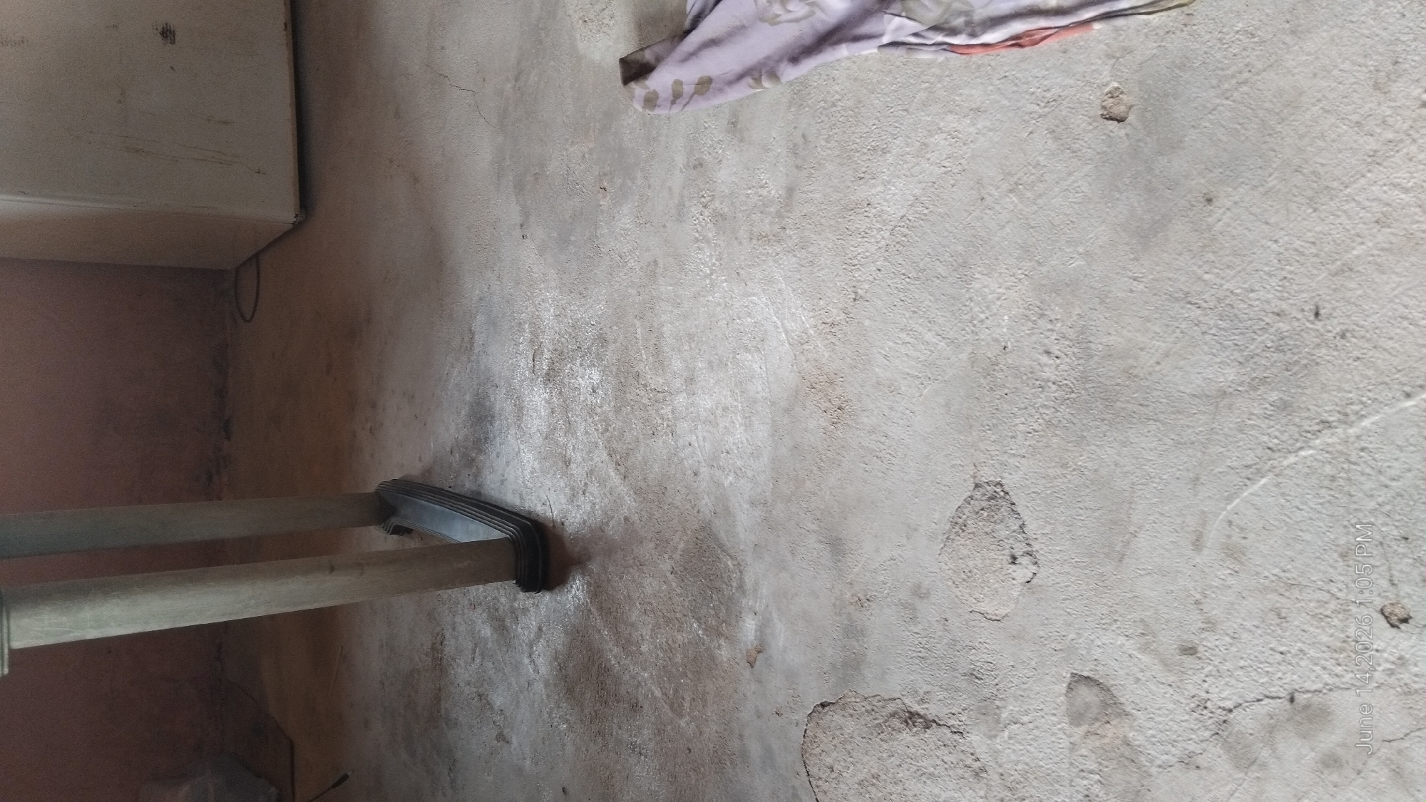 The image depicts a close-up view of a concrete floor in what appears to be an unfinished or storage area, possibly located in Howayi, Nigeria. The floor is uneven and shows signs of wear, with several patches of discoloration and what may be repairs or imperfections in the concrete. The lighting is dim, casting shadows from a vertical object on the left, which is partially visible and appears to be a table or furniture leg made of a light-colored material with a dark base. In the background, a portion of a large, white appliance, possibly a refrigerator, is visible. On the right side of the image, a piece of fabric, possibly a blanket or towel with a floral pattern in shades of purple and red, is draped over the edge of the frame. The overall impression is one of a utilitarian space with minimal furnishing and some signs of use or neglect.