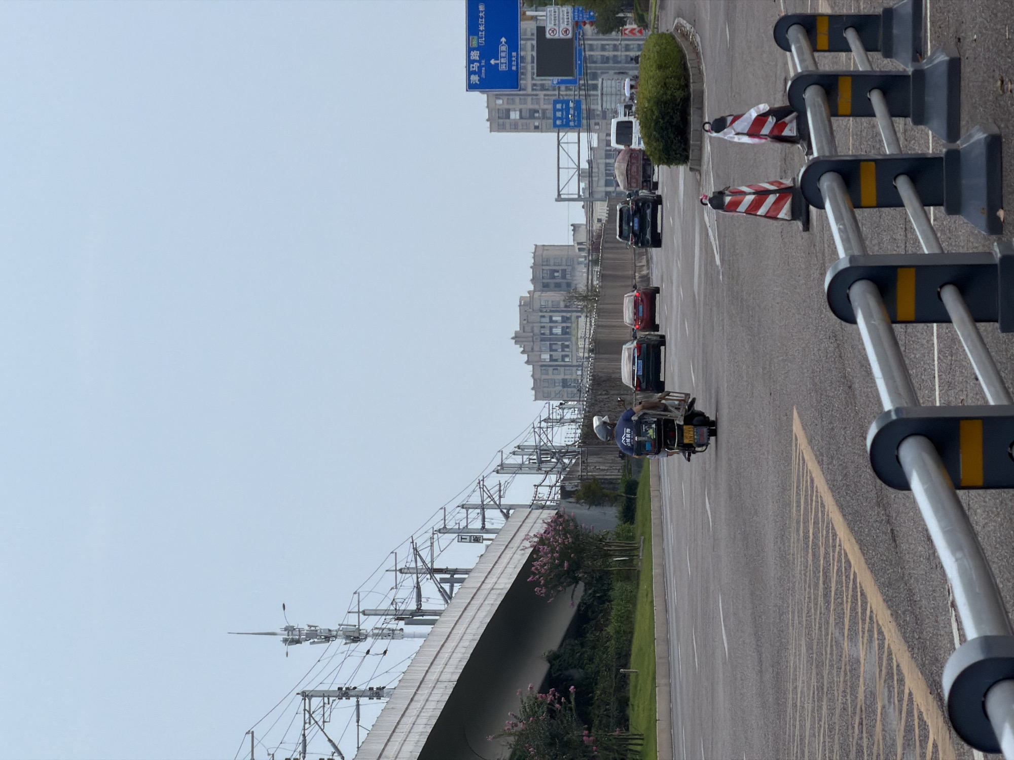 The image depicts a street scene in Jiangjin, China, on an overcast day. In the foreground, a metal railing with dark grey posts and silver rails separates the viewer from the road. Yellow and black markings are visible on the posts and on the road surface, including diagonal yellow lines and solid white lane markers.
Several vehicles are present on the road. A person on a scooter, wearing a dark blue shirt and a helmet, is traveling away from the camera in the left lane. The scooter has a large box attached to the back. Further down the road, several cars are visible: a black sedan, a red sedan, a black SUV, a truck with a covered load, and a white van. Orange and white traffic cones, some with reflective tape, are placed near the road's edge.
In the background, a large elevated concrete structure, likely an overpass or bridge, dominates the left side of the frame. Electrical wires and utility poles are suspended beneath and alongside this structure. Trees with pink flowers are planted near the base of the overpass. Beyond the road and overpass, buildings of various sizes and architectural styles can be seen, including several multi-story apartment buildings. A blue road sign with Chinese characters and directional arrows is positioned on the right side of the road, indicating directions to "津马路" (Jinma Road) and "长江大桥" (Yangtze River Bridge).