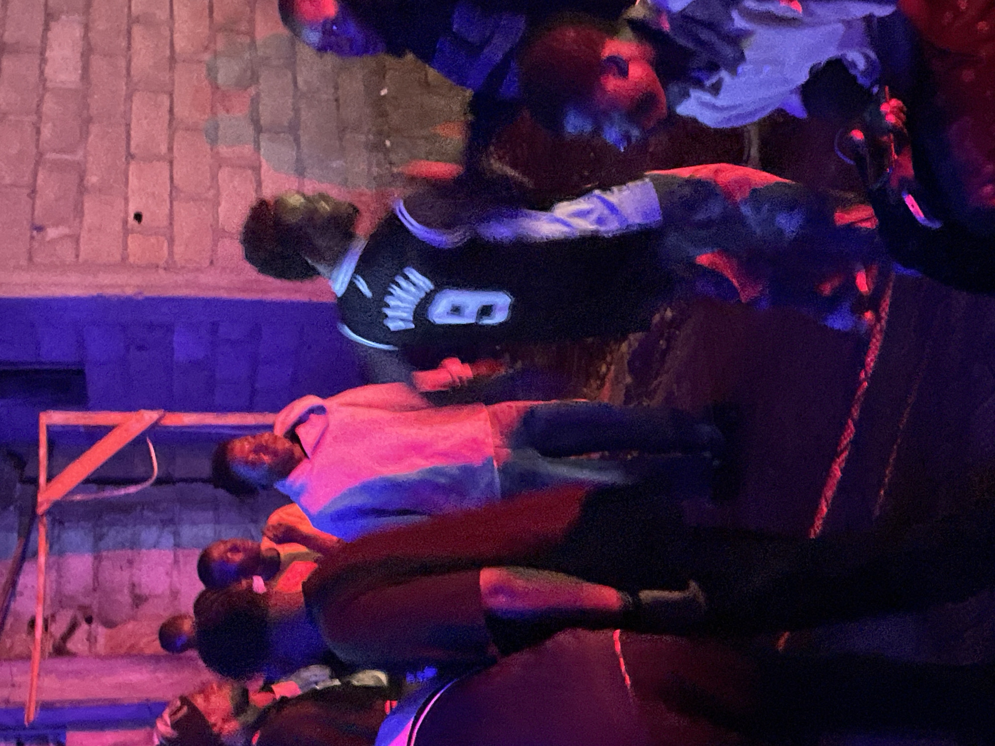 The image depicts an outdoor gathering of several individuals, predominantly young men, in a dimly lit environment illuminated by red and blue lights. The background features a brick wall and what appears to be makeshift wooden scaffolding, suggesting a potentially informal or construction-adjacent setting. The individuals are dressed in casual attire, including t-shirts, hoodies, and what seems to be a basketball jersey. Some individuals are facing away from the camera, while others are looking towards it or interacting amongst themselves. The overall atmosphere suggests a social event or a casual assembly. The specific location in the city of Durumi, Nigeria, is not explicitly identifiable from the visual information provided in the image itself.