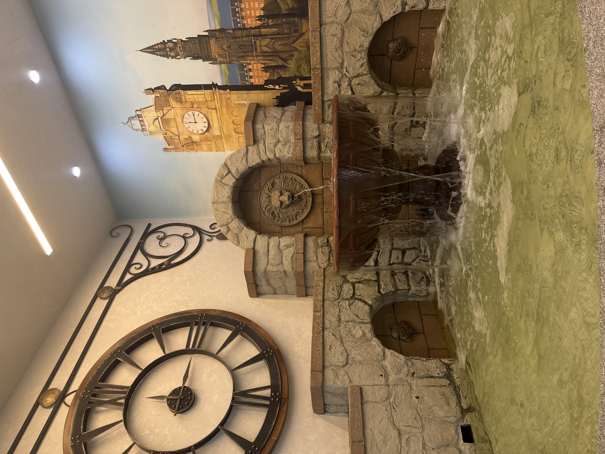 The image displays an indoor decorative fountain integrated into a wall mural. The mural depicts a city skyline with prominent clock towers, suggestive of London, against a pale blue sky. The fountain itself is constructed to resemble aged stone with arched openings. Water cascades from a lion's head spout, flowing into a shallow pool below. Adjacent to the fountain, on the textured wall, is a large, ornate Roman numeral clock with dark metal hands. The overall aesthetic combines architectural elements with natural water features, creating a unique visual display. The scene appears to be indoors, possibly in a public space or a creatively designed private residence, given the lighting and the nature of the installation. There are no people visible, and the mood is tranquil, focused on the gentle movement of water and the decorative artistry. The lighting suggests it is daytime or that artificial lighting is used to illuminate the area. No text is visible within the image. The context provided indicates this is located in Yekaterinburg, Russia.