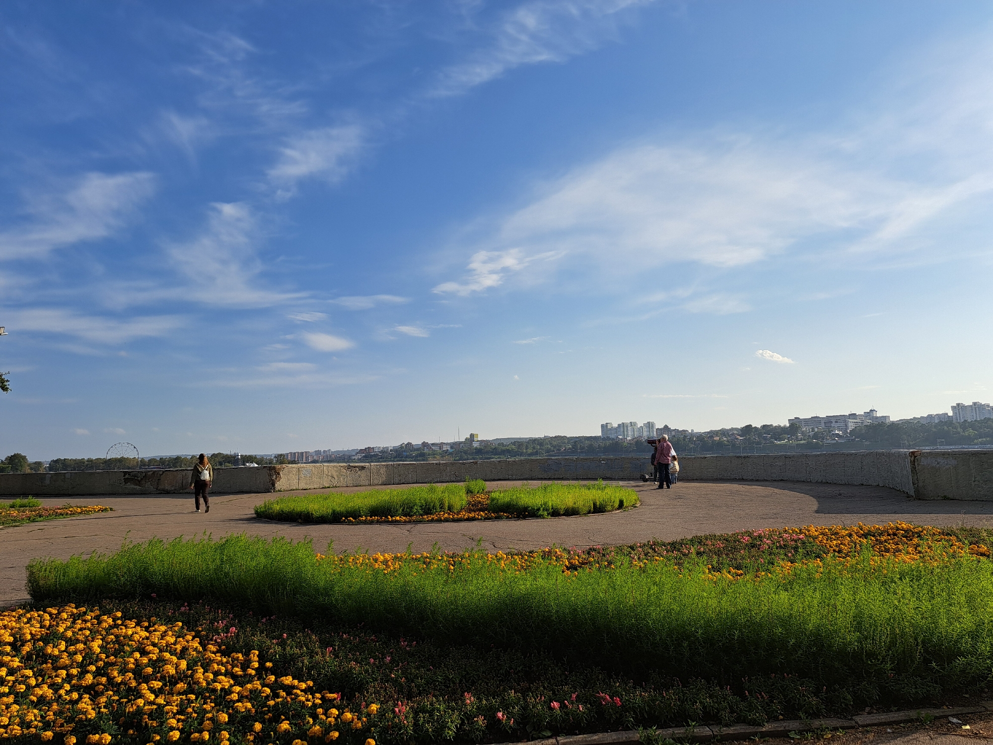 The image depicts a park area with cultivated flower beds and a walkway, situated along a waterfront with a cityscape in the background. The foreground is dominated by vibrant yellow marigolds and lush green plants, arranged in distinct beds. A paved walkway curves through the park, bordered by concrete barriers. In the middle ground, two individuals are visible. One person, wearing a light-colored backpack and dark clothing, walks away from the camera. Further to the right, another person in a pink shirt and dark pants appears to be interacting with a scooter or a similar wheeled device, with another individual partially visible behind them.
The background reveals a wide river or body of water, beyond which lies a city skyline with numerous buildings of varying heights, suggesting an urban environment. A Ferris wheel is faintly visible in the distance on the left side of the frame, a common landmark in city parks. The sky is a clear blue, with scattered wispy white clouds. The lighting suggests it is daytime, possibly late afternoon, given the angle of the sunlight casting subtle shadows.
This scene is characteristic of the Lower Angara Embankment in Irkutsk, Russia, specifically near the Icebreaker Monument, which is often adorned with flowerbeds and offers views of the Angara River and the city's panorama. The presence of the Ferris wheel further supports this identification, as one is located in the immediate vicinity.