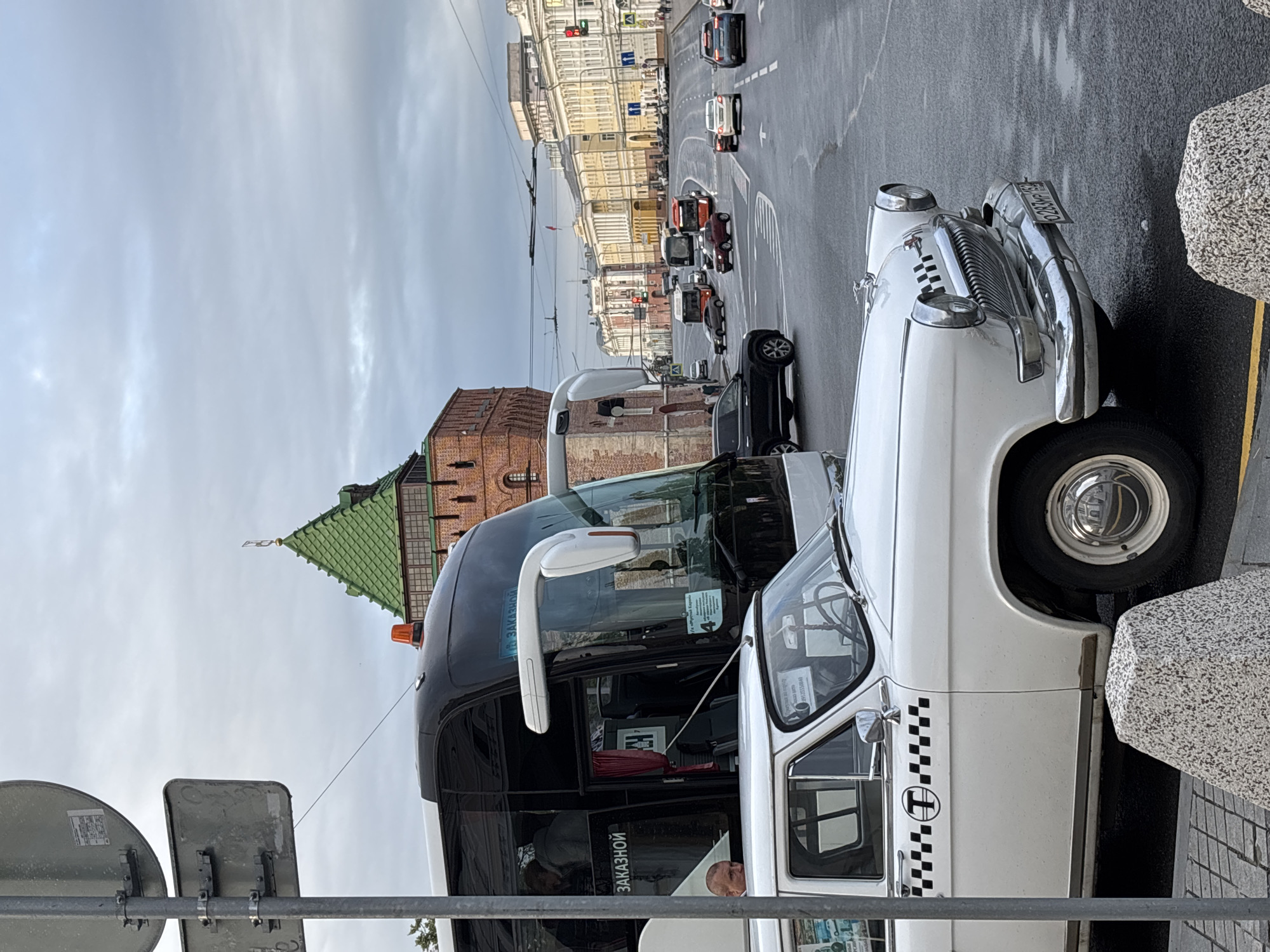 This is a street-level, eye-level medium shot of a street in Nizhny Novgorod, Russia. The sky is overcast and grey. In the foreground, a white classic car, possibly a GAZ-24 Volga, is parked. It has a checkered taxi pattern on the driver's side door with the letter "T" inside a circle. The car's front bumper and grille are visible, as is a license plate that appears to read "0283MP 152". Behind the classic car, a black and white bus is parked, with "ЗАКАЗНОЙ" (ordered) written on its side. The bus has an orange flashing light on its roof.
In the background, a tall brick tower with a green roof is visible, which is likely part of the Nizhny Novgorod Kremlin. Further down the street, there are multi-story buildings with classical architecture, some painted in yellow and white. Several other vehicles, including a red SUV and other cars and buses, are present on the wet asphalt road, suggesting recent rainfall. Traffic lights are visible at an intersection in the distance. The overall scene depicts an urban environment with a mix of historical and modern elements.