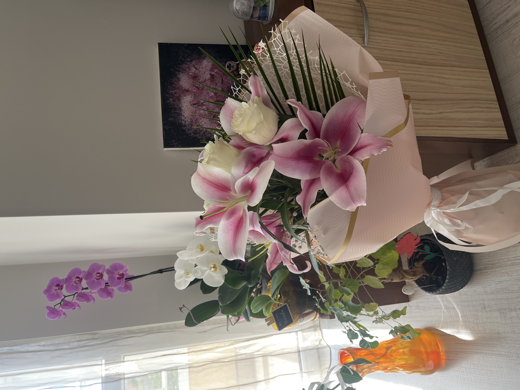 The image depicts a bouquet of flowers, predominantly pink lilies and white roses, wrapped in light pink paper with a white mesh overlay. The bouquet is placed on a surface, likely a table, near a window with beige blinds. An orchid with purple and white blossoms is also visible. An orange vase stands nearby. A framed abstract painting hangs on the wall in the background, and a wooden cabinet or shelf is situated to the right of the bouquet. There's no verifiable indication of the location being Blagoevgrad, Bulgaria, in the image.
