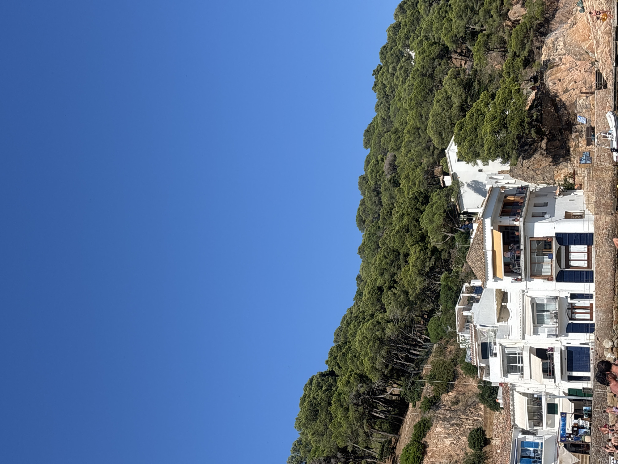 The image shows a view of the coastline in the municipality of Palafrugell, Spain, specifically focusing on the village of Tamariu. In the foreground, a rocky shoreline is visible with a few people sunbathing and walking. A small white boat is docked at the water's edge. Behind the shoreline are a series of white buildings with multiple stories, some featuring balconies and awnings. These buildings appear to be houses or small hotels, typical of a coastal Mediterranean town. The buildings are situated on a hillside that rises sharply behind them. This hillside is densely covered with green pine trees, characteristic of the Mediterranean landscape. The sky above is a clear, deep blue. The scene suggests a sunny day in a popular tourist destination.