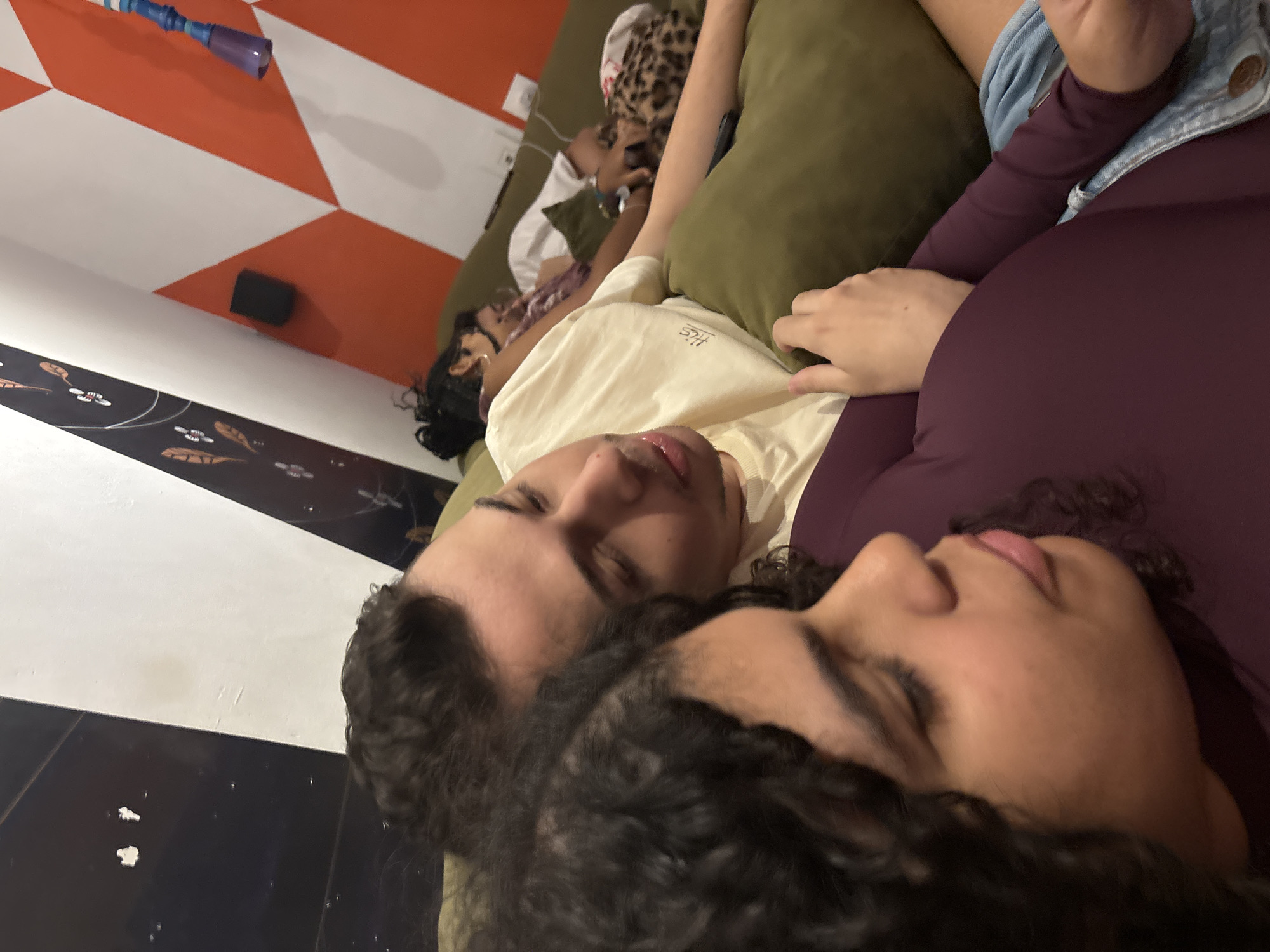 A medium shot captures two dark-haired individuals lying side-by-side on a green cushion, seemingly indoors. The individual on the left is wearing a pale yellow t-shirt, while the one on the right is clad in a dark purple, form-fitting long-sleeved shirt, also sporting a denim jacket. In the background, two more people are visible, appearing to be resting on the same cushion. The foreground floor shows a design of black and white, while the background wall features a geometric pattern of orange and white. A small, black, rectangular object is attached to the wall, and a blue and purple object is on the orange and white patterned wall. While the image provides clear details of the immediate scene, specific location details within São Paulo, Brazil, are not discernible.