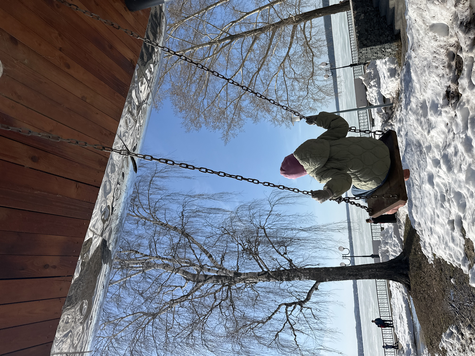 A low-angle, landscape-oriented photograph shows a swing suspended from chains in what appears to be a public park in Votkinsk, Russia, during winter. A child wearing a pink hat and a light green, quilted jacket is sitting on the wooden swing, facing away from the camera. The ground around the swing is covered in snow, and the adjacent structure to the left features brown wooden boards and a shiny, reflective vertical panel. Above the swing, bare tree branches stretch across a clear, blue sky. In the distance, across the frozen water (likely the Votkinsk reservoir), a metal fence and light posts are visible. Two or three people, rendered small due to the distance, are standing near the water's edge. The whole image has a slight tilt, angled to the left.