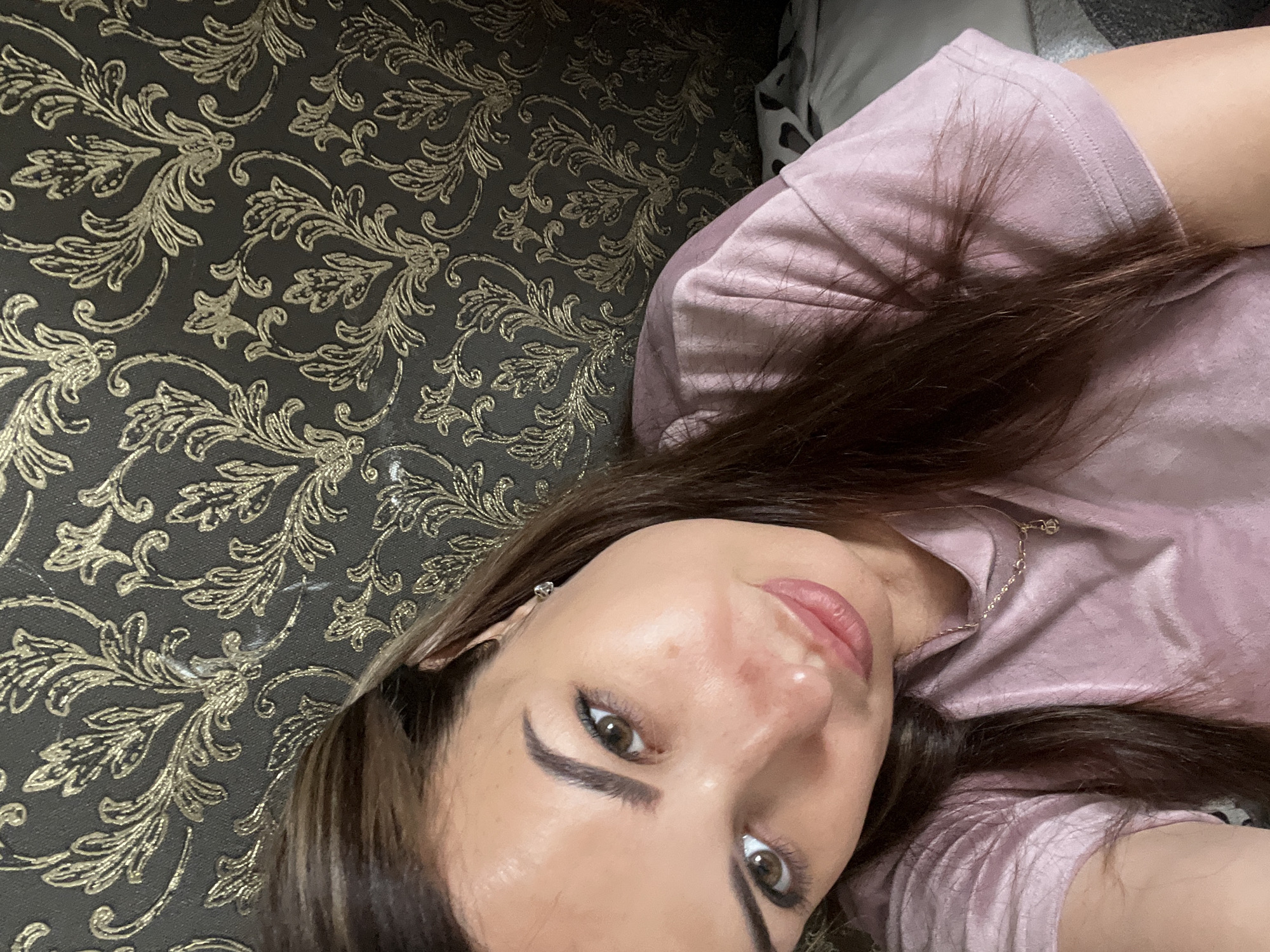 A woman with long brown hair and light brown eyes is pictured from the chest up. She is looking at the camera with a slight smile. She is wearing a light pink t-shirt and a delicate gold necklace. The background is a dark, textured wallpaper with a gold damask pattern. The lighting is soft and natural. The image appears to be a selfie, taken indoors.