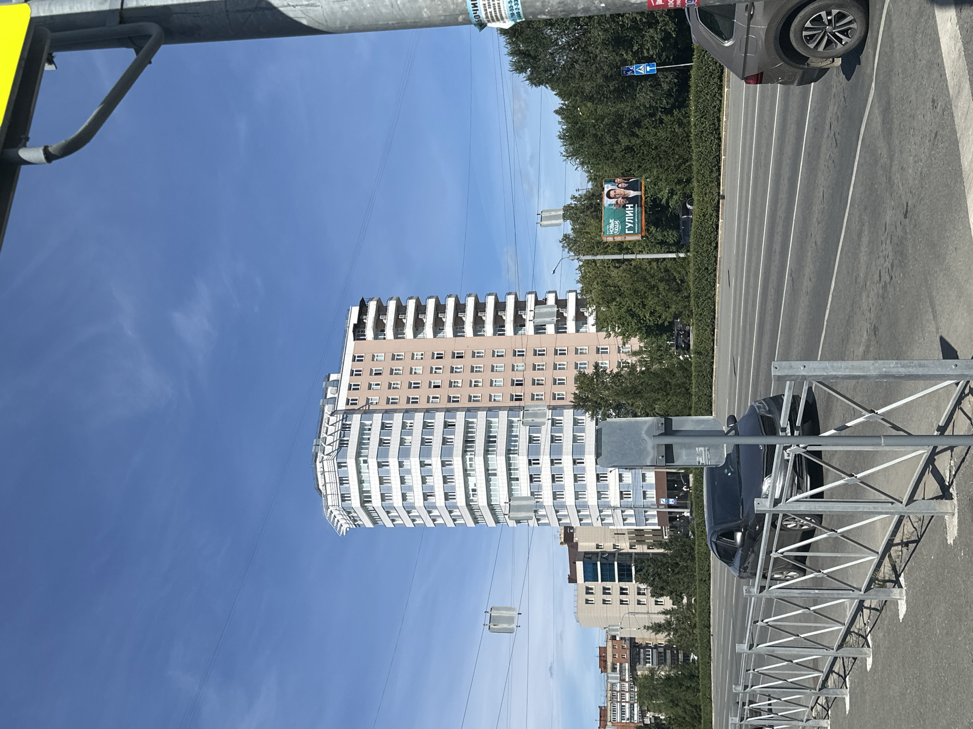 The image depicts a street scene in a city under a clear blue sky. In the foreground, a metal fence with a criss-cross pattern runs along the sidewalk. A grey car is visible on the road to the right, and another dark grey sedan is on the road to the left, approaching the viewer.
Dominating the middle ground is a tall, modern apartment building with a white, blue, and beige facade, featuring numerous windows. To the left of this building, other residential structures of varying heights and architectural styles are visible, some with balconies. Trees line the road, creating a green buffer between the buildings and the street.
In the background, a billboard with images of people and Russian text, including "ГУЛИН", is placed among the trees. A lamppost and traffic signs are also present in the scene. Utility wires are strung across the sky, and a square grey object is suspended from them. The overall environment suggests an urban setting on a sunny day.