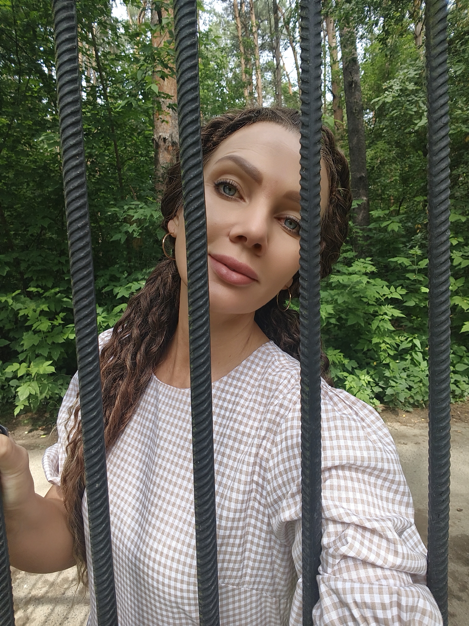 A woman with brown, curly hair and light blue eyes is pictured in a park-like setting, peering through black metal bars. She is wearing a light-colored, checkered dress with puffy sleeves. The background is filled with lush green foliage and trees. The woman's left hand is visible, resting on the bars.