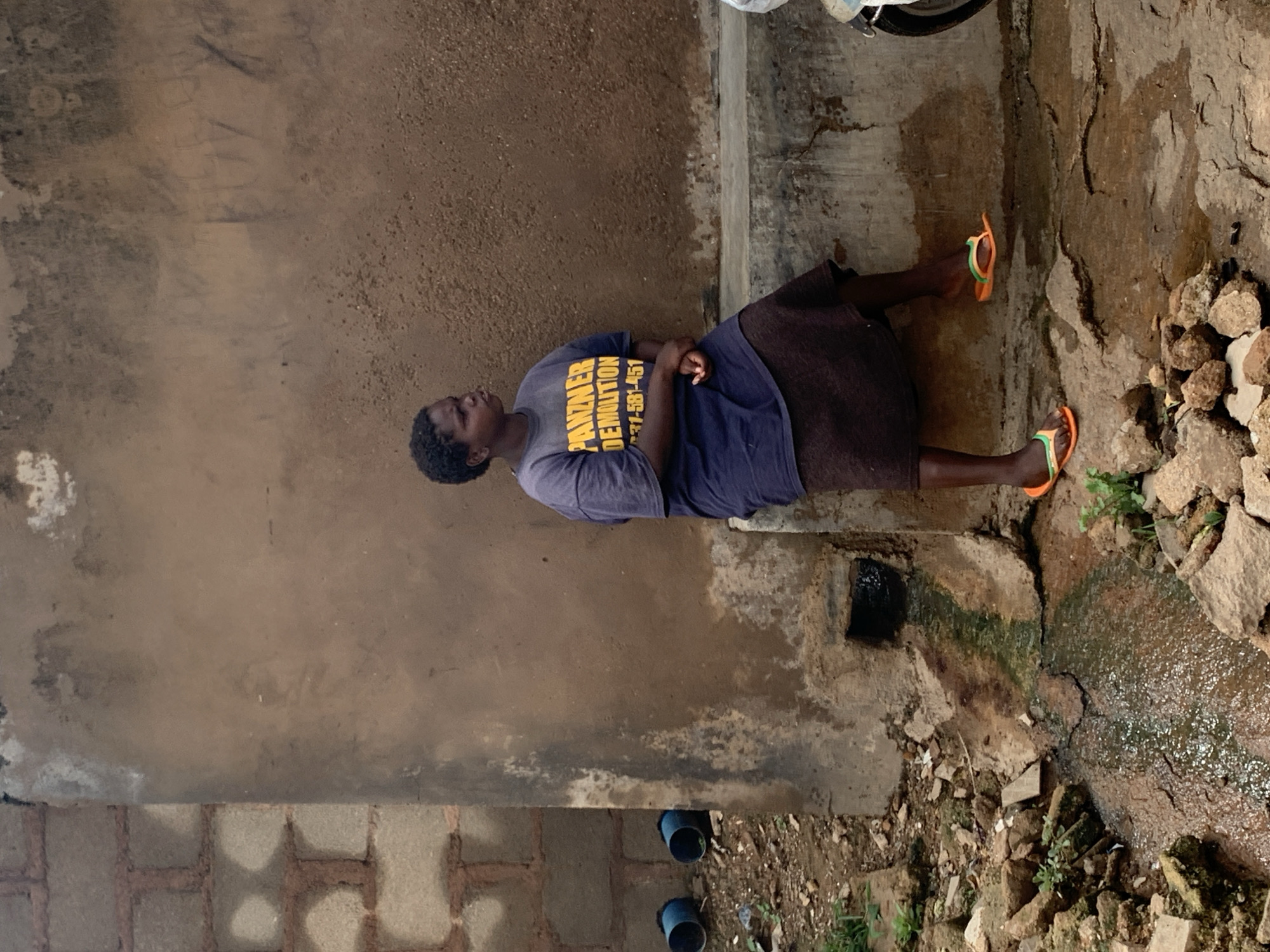 The image shows a person lying down on the ground outdoors. The person is wearing a blue t-shirt with yellow text and a dark skirt. They are also wearing orange and green flip-flops. The ground appears to be a mix of dirt, concrete, and rocks. There is a drain or pipe visible in the lower right portion of the image, and some green plants growing around it. In the background, there is a textured wall.
The text on the person's shirt is partially visible and appears to read "PANZNER DEMOLITION" followed by a phone number that seems to end in "-581-451".
The scene suggests a casual or perhaps resting pose. The lighting appears to be natural, possibly indicating daytime. The ground and surrounding environment suggest an outdoor, possibly somewhat undeveloped, area. The presence of the drain and rough terrain could indicate an industrial or construction-adjacent location. The overall impression is of a person taking a moment to rest in a somewhat utilitarian environment.