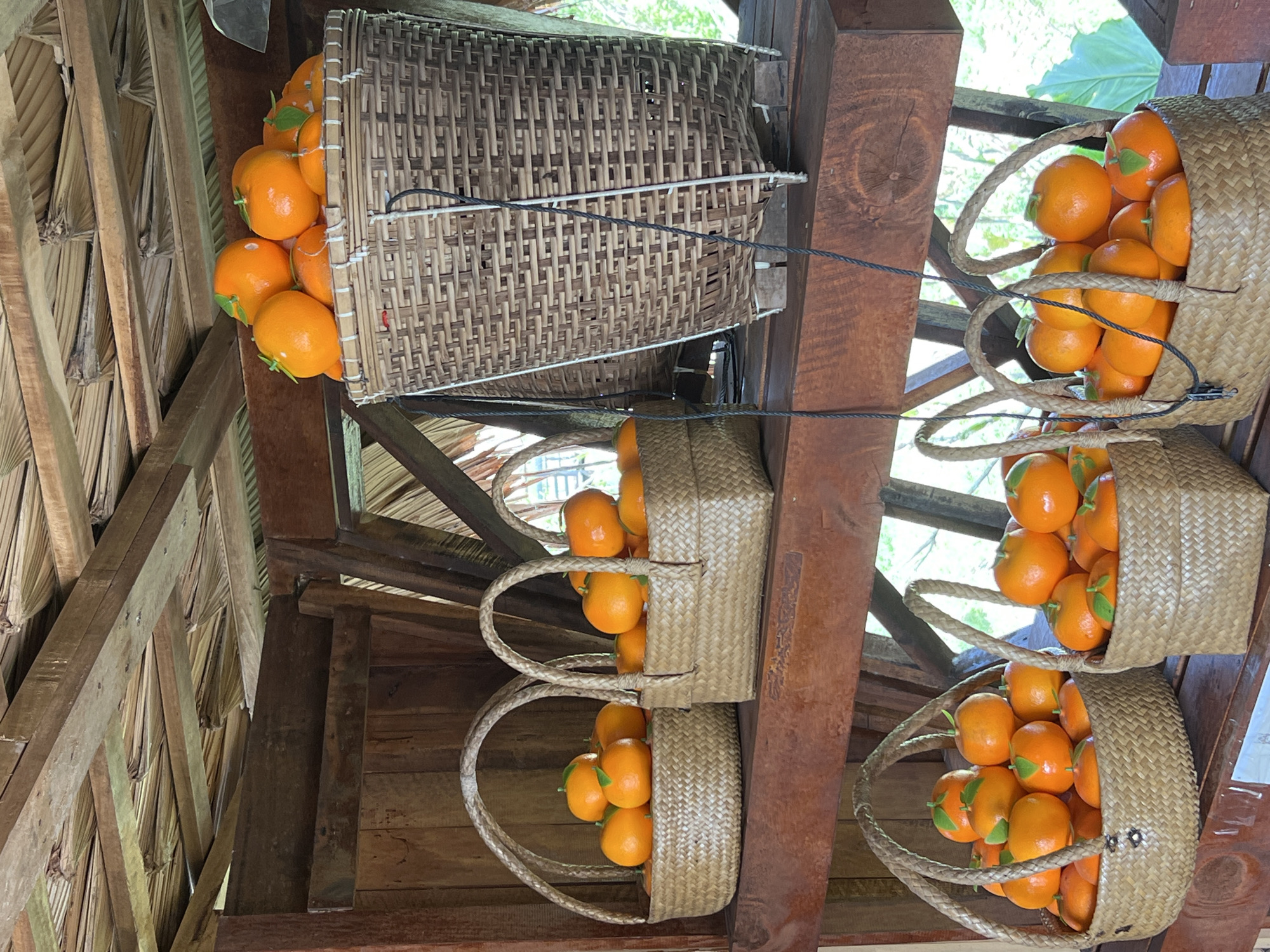 This media file shows a close-up view of several woven baskets filled with bright orange fruits, likely tangerines or small oranges. The baskets are arranged vertically, with one large basket at the top and two smaller baskets stacked below it. The large basket is filled to the brim with the fruit, and strings are attached to its sides, suggesting it may be suspended. The two smaller baskets are also full, with one basket resting on top of the other. The handles of the smaller baskets are visible, made of braided material.
The setting appears to be rustic and outdoors, possibly a market stall or a fruit stand. Wooden beams and palm fronds are visible in the background, indicating a tropical or subtropical location. The lighting suggests it is daytime, with natural light illuminating the scene. There are no people visible in the media file. The overall impression is one of abundance and freshness. The location context provided is Tambon Pa Kae Bo Hin, Thailand, which aligns with the visual cues of a tropical setting. No visible text is present in the media file.