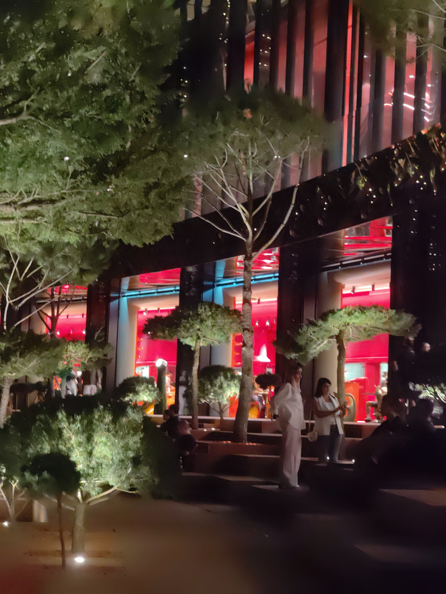 The image depicts an outdoor scene at night, with a modern building in the background featuring red and blue lighting. In the foreground, several people are visible, some standing and some seated, amidst strategically placed trees and bushes. One woman in the foreground is wearing a white outfit, while another woman in a white vest stands next to her. The ground appears to be a sandy or gravel path, with some artificial lighting illuminating the area. The overall impression is of a well-lit urban space, possibly a plaza or courtyard. The location in Krasnodar, Russia is not visually discernible from the image content.