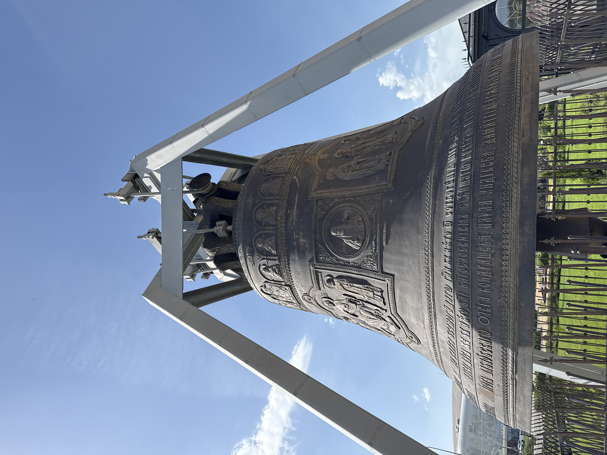 The image shows the Tsar Bell, a large bronze bell located in Nizhny Novgorod, Russia. The bell is mounted on a white steel frame. The bell has intricate carvings and inscriptions in Cyrillic script on its surface. It is situated outdoors, with a blue sky and some clouds visible in the background. A decorative metal fence surrounds the bell. Green grass is visible behind the fence.