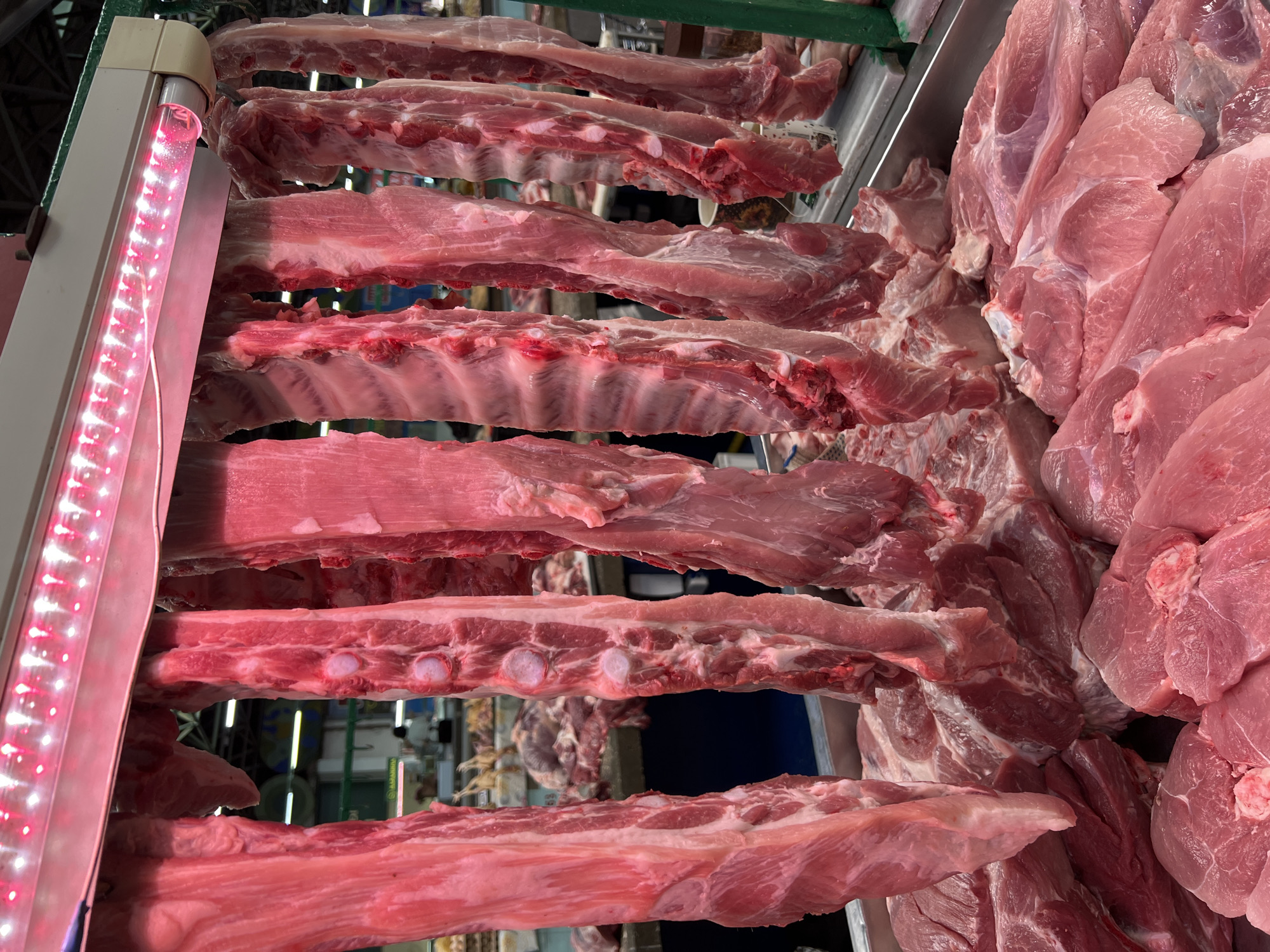 This image depicts a raw meat display within an indoor market in Krasnodar, Russia. The primary subjects are numerous cuts of fresh pork, both hanging and displayed on a counter.
In the upper midground, several large, elongated cuts of raw pork, appearing to be bone-in ribs and loins, hang vertically from hooks. These cuts exhibit a reddish-pink coloration with visible muscle tissue, fat, and bone structures, including prominent vertebral sections in some pieces. Directly above these hanging meats is a horizontal light fixture containing a row of active red LED bulbs, casting a distinct reddish glow onto the meat below. The fixture is supported by a light-colored frame.
Below the hanging meat, a significant quantity of raw pork cuts is arranged in a metallic or stainless steel display tray. These cuts are predominantly boneless portions, such as chops or stewing meat, stacked and spread across the surface. They display a light pink to reddish-pink color, consistent with fresh pork.
In the background, the scene expands into a bustling market environment. Blurred figures of individuals, likely market vendors and customers, are visible among other stalls and displays. One person in a light-colored top is discernible further back. The overall background suggests an expansive, active commercial setting with overhead structures and ambient lighting distinct from the specialized red light over the meat. No specific actions or interactions are clearly detailed due to the distance and blurriness of background figures.