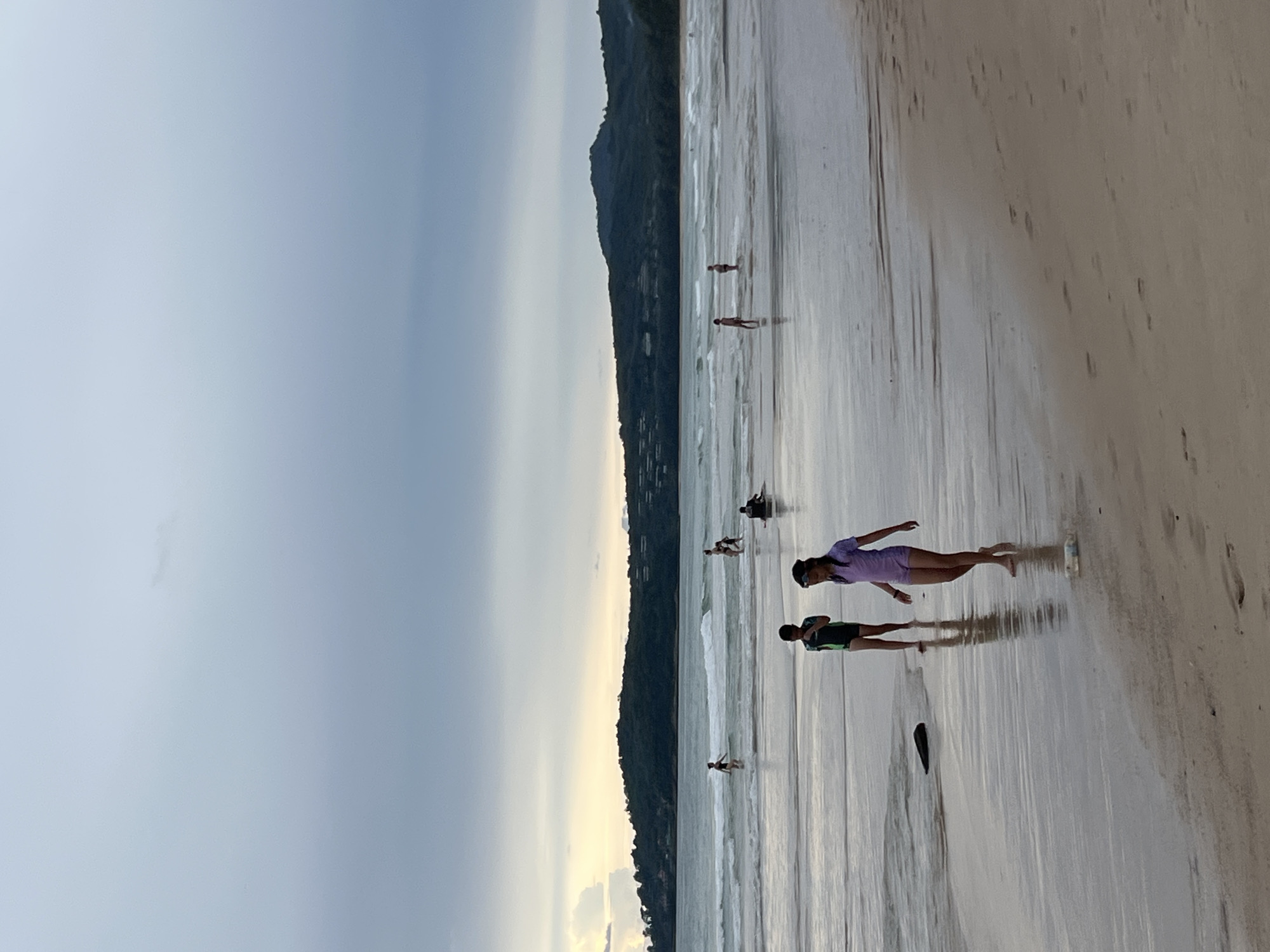 A group of people are on a beach in Choeng Thale, Thailand. The image is oriented vertically, with the ocean on the left and the sand on the right. The sky is overcast, and the sun appears to be setting, casting a warm glow on the horizon. The water is calm with gentle waves rolling in.
Several individuals are visible on the wet sand, some walking, others standing. In the foreground, a young girl in a purple shirt and dark shorts is posing, with her reflection visible in the wet sand. Beside her, a boy in a green and black shirt and dark shorts is also standing. Further out in the water, more people can be seen scattered across the beach.
In the background, a lush, green hillside rises steeply from the coastline, suggesting a tropical location. The hillside is dotted with buildings, indicating a populated area. The overall mood of the image is tranquil and relaxed, with the soft lighting and peaceful setting contributing to a sense of calm. There are no visible text elements in the image.