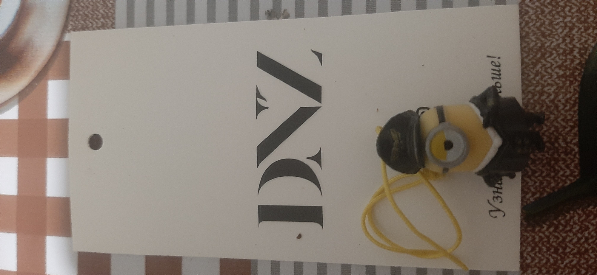 A white tag with black lettering displays the word "DNZ" in large, ornate letters. Below the lettering, a small Minion toy is attached to the tag by a yellow string. The Minion is wearing a black aviator hat and a dark suit. The tag is placed on a surface with a checkered pattern. Text below the Minion reads "Узнать больше!" which translates to "Learn more!". The background shows a plaid tablecloth. The image is a close-up shot, with no significant environmental context. No specific location in Yessentukskaya, Russia, is identifiable from the visual information.