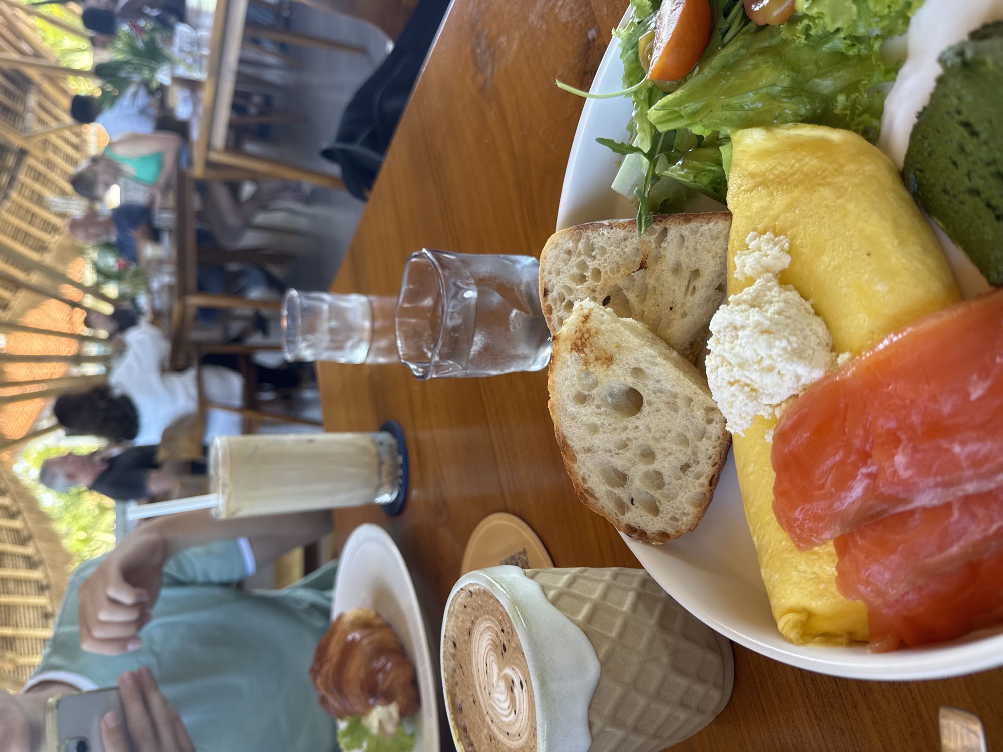 Captured at an elevated angle, the photograph shows a table at a restaurant in an unidentified city. The table is wooden. Various food and beverage items are set on the table. A plate is filled with an omelet, slices of bread, salmon, cheese, salad and a green slice of food. A cup of coffee and a croissant are also placed nearby. Clear glasses of water and iced coffee are also seen on the table. Several people are visible in the blurry background. A person is taking a picture of the food. The location appears to be a restaurant with bamboo structures.