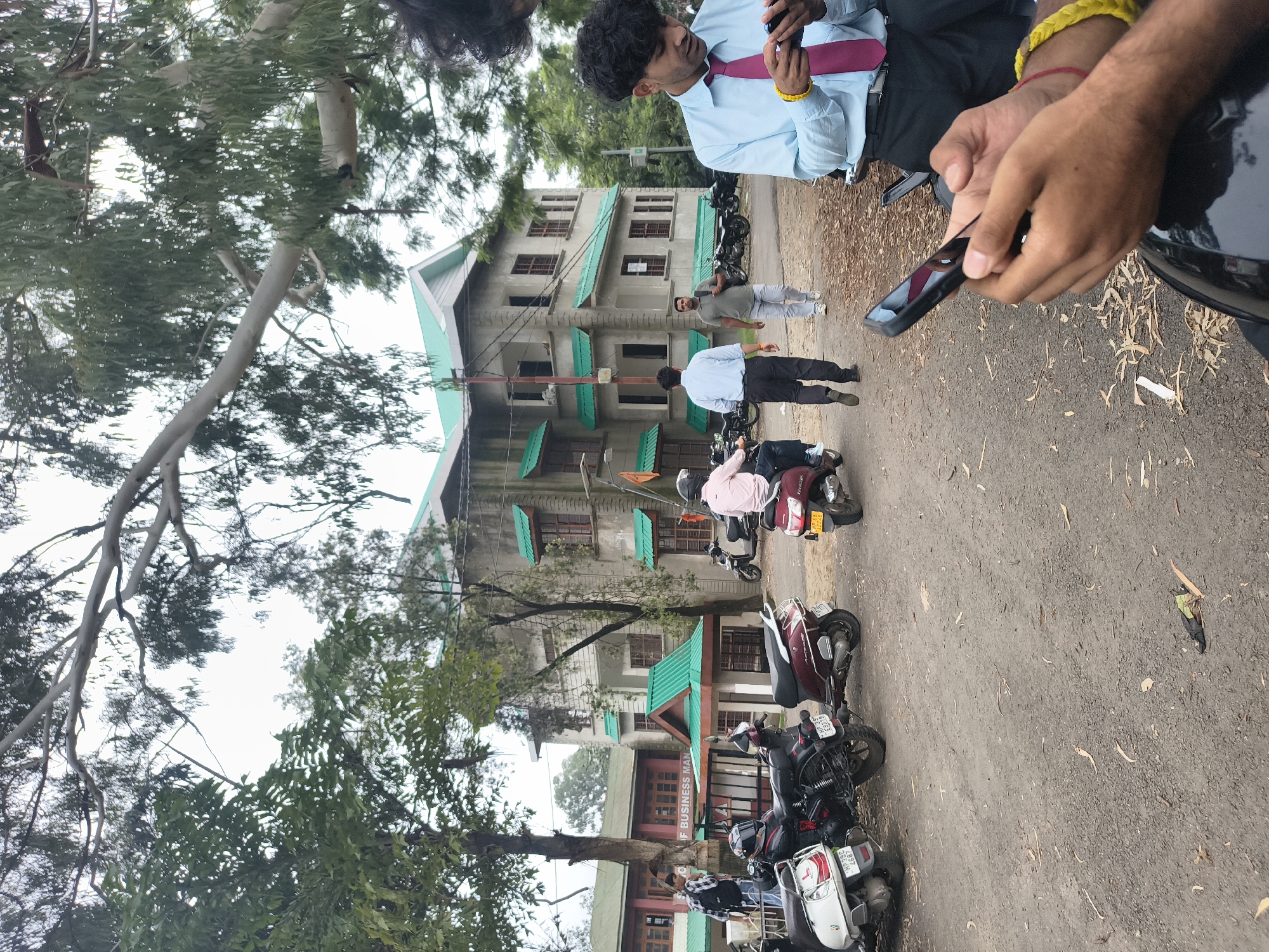 The image depicts a scene outside a building that appears to be an educational institution, possibly the Himachal Pradesh University Business School, located in Dharamshala, India. The architecture features multiple stories with a light-colored exterior and distinctive green sloping roofs. Several people are present, some on scooters and others walking. In the foreground, a man wearing a blue shirt and a pink tie is looking at his mobile phone, with his hands visible. He has yellow and red wristbands. To his left, another man in a pink shirt is riding a maroon scooter. Behind him, two men in formal attire are walking away from the camera. One is in a light blue shirt and dark pants, and the other is in a gray t-shirt and light-colored pants, carrying a backpack. Several parked scooters and motorcycles are visible on the gravelly ground. The environment is characterized by trees with dense foliage, and the sky appears overcast. A flag with an orange symbol is attached to the side of the building.