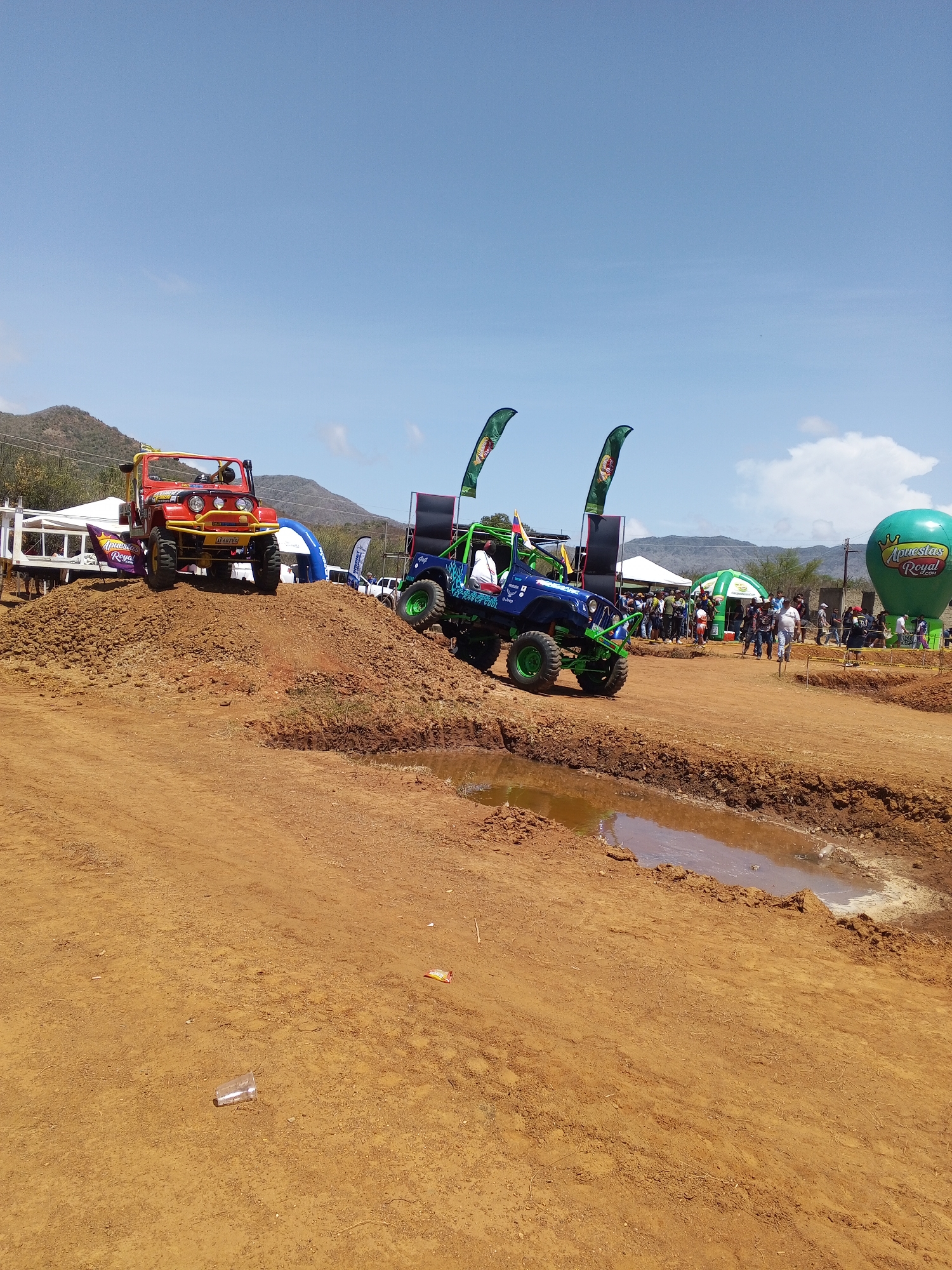 The image shows an off-road vehicle event in an unidentified location. In the foreground, there is a muddy dirt track with a puddle of water. Two modified Jeeps are positioned on dirt mounds; one is red and yellow, and the other is blue and green. There are speakers and flags with the “Apuestas Royal” logo beside the blue and green Jeep. A green “Apuestas Royal” balloon is visible in the background to the right. A crowd of people can be seen standing in the background. Mountains and a partially cloudy sky are in the distance. A plastic cup sits discarded in the foreground dirt.
