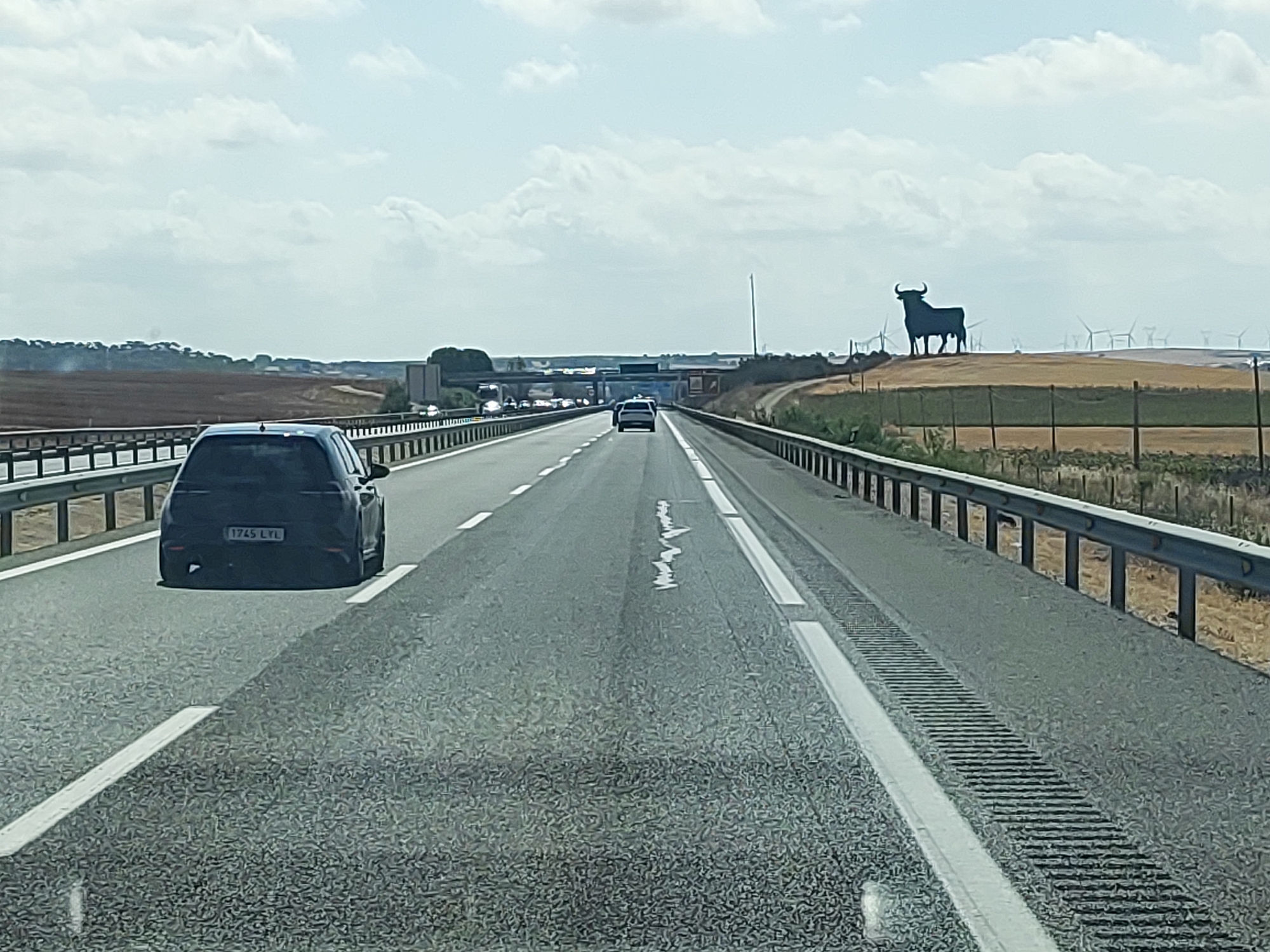 The image shows a view from a vehicle driving on a highway in Spain, likely near Castillo de Garcimuñoz, as evidenced by the iconic Osborne bull silhouette visible on a distant hill. The highway is paved with asphalt and marked with white lines. Guardrails line the sides of the road. In the foreground, a black car with the license plate "1745 LYL" is visible. Further down the highway, other vehicles are present, including a white SUV. The landscape consists of dry, grassy fields and some vegetation, with wind turbines scattered in the distance. The sky is cloudy.