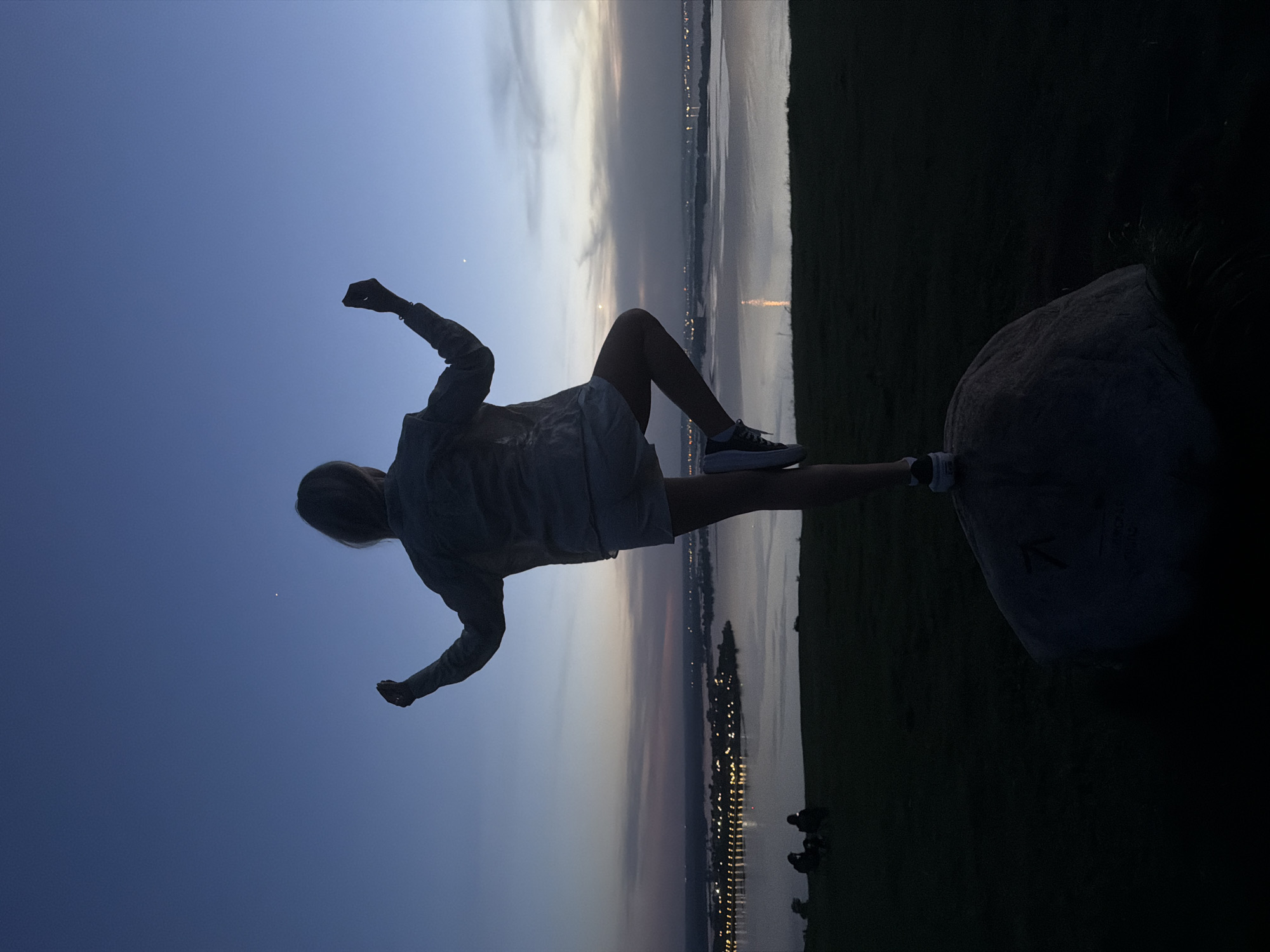 A person is sitting on a rock in Innopolis, Russia, with their legs extended towards the city lights reflecting on the water at dusk. The person is wearing shorts, a jacket, and sneakers, and is silhouetted against the sky. The city lights are visible across the water. The environment includes grass and a rock. The sky is transitioning from blue to light orange where the sun sets.
