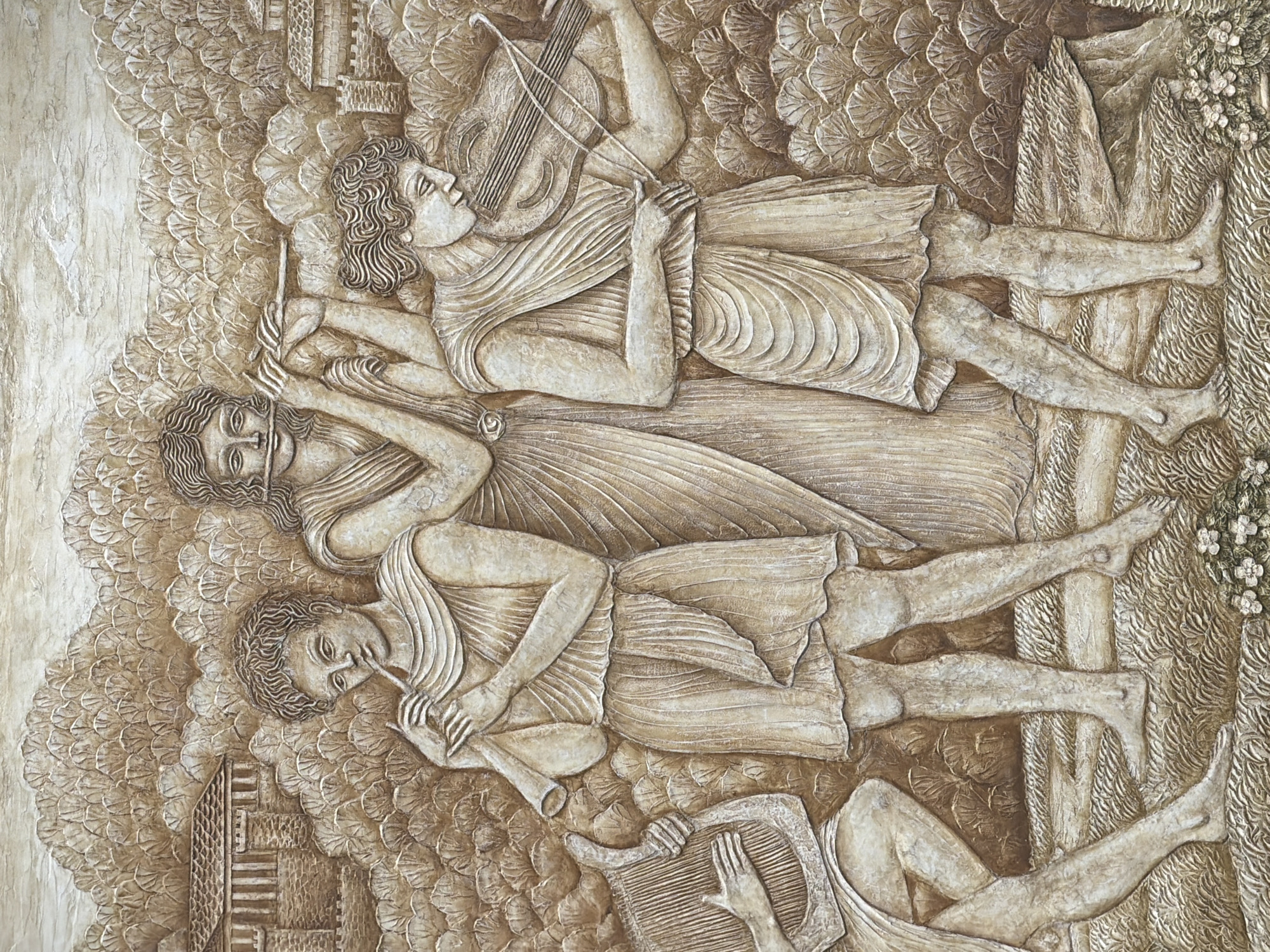 This is a relief sculpture depicting four figures playing musical instruments in an outdoor setting. The figures are rendered in a classical style, wearing draped clothing. One figure is playing a violin, another a flute or pipe, and a third a harp-like instrument. The fourth figure's instrument is partially obscured. The background includes stylized trees and foliage, along with a representation of a building or structure. The overall texture of the relief suggests it's made of stone or a similar material. The image does not contain information pertaining to the city of Sochi, Russia.