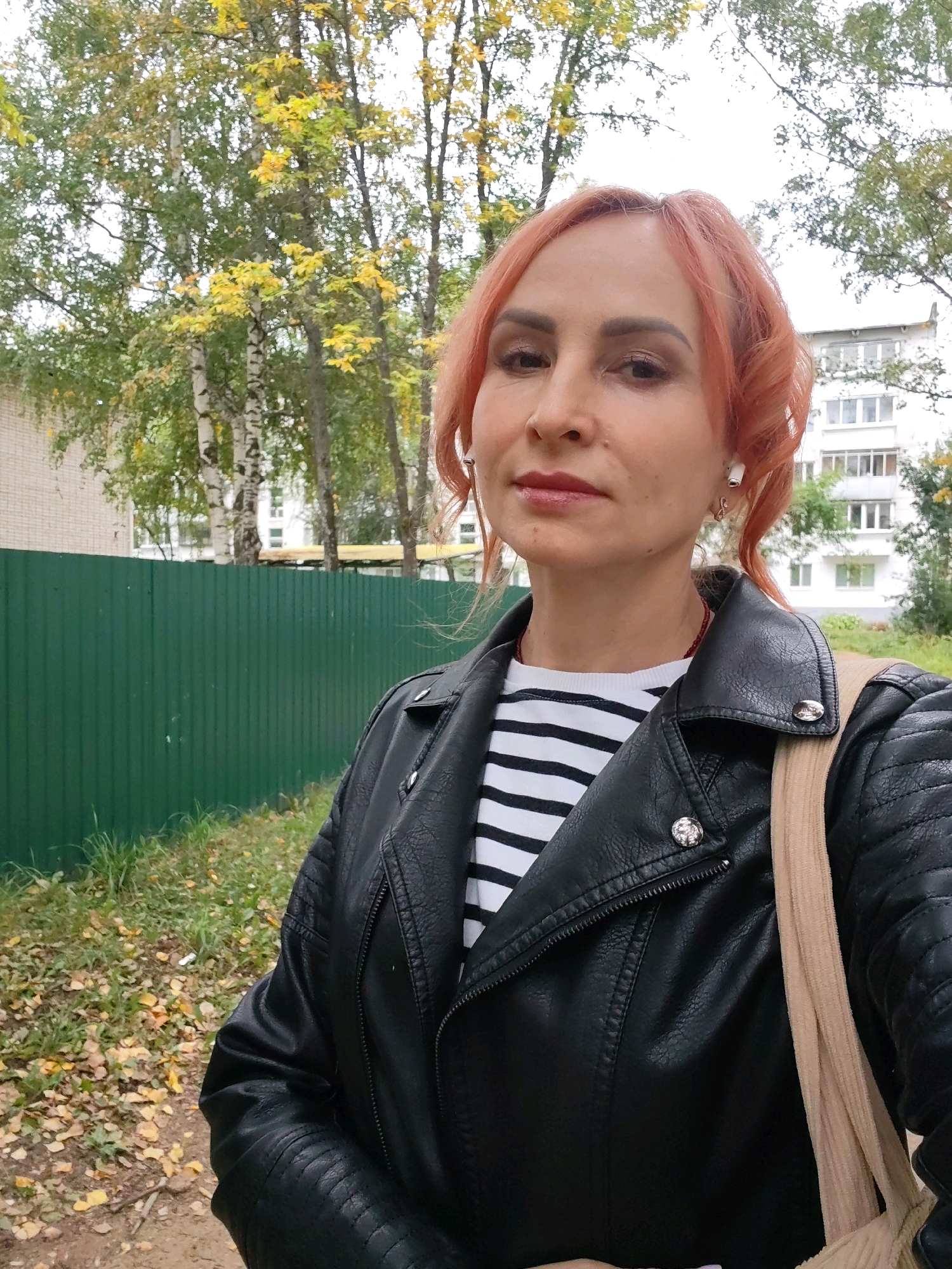 An outdoor medium shot shows a woman with reddish-pink hair and makeup, wearing a black leather jacket over a striped shirt and white earbuds. She is standing in front of a green metal fence with a building and trees visible in the background. The scene appears to be in a residential area, possibly a courtyard, with fallen leaves on the ground, suggesting it is autumn. The image is a selfie taken by the woman. The location is the city of Chaikovsky, Russia.