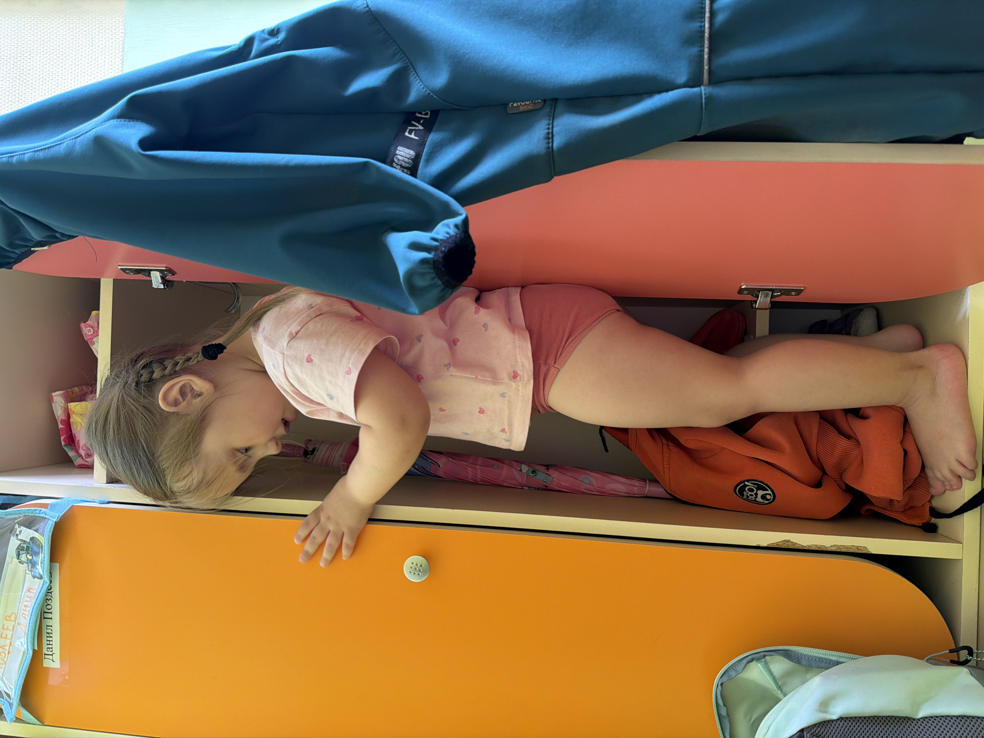 A young girl with light brown hair, styled in braids with black elastics, is depicted lying horizontally inside an open shelf of a colorful cabinet. She is wearing a light pink short-sleeved t-shirt with small heart patterns and pink underwear. Her body is stretched out within the shelf, and she appears to be looking downwards or to her side with a curious and playful expression.
The cabinet features multiple levels with sections in peach/coral and orange colors. Above the girl, a blue jacket hangs, displaying text that includes "FV-B" and "Favourite LAND". Inside the shelf with the girl, a pink umbrella and an orange sweatshirt with a white circular logo are visible towards her feet. On the shelf below, an orange drawer is partially visible, and to its left, a light blue bag or folder with Cyrillic text, including "Данил Позд-" and "ОЛЕЕВ ДАНИЛ", is present. A grey and light green backpack is also partly seen at the bottom right. The scene takes place indoors, suggesting a home environment in Chaikovsky, Russia, likely during the daytime.