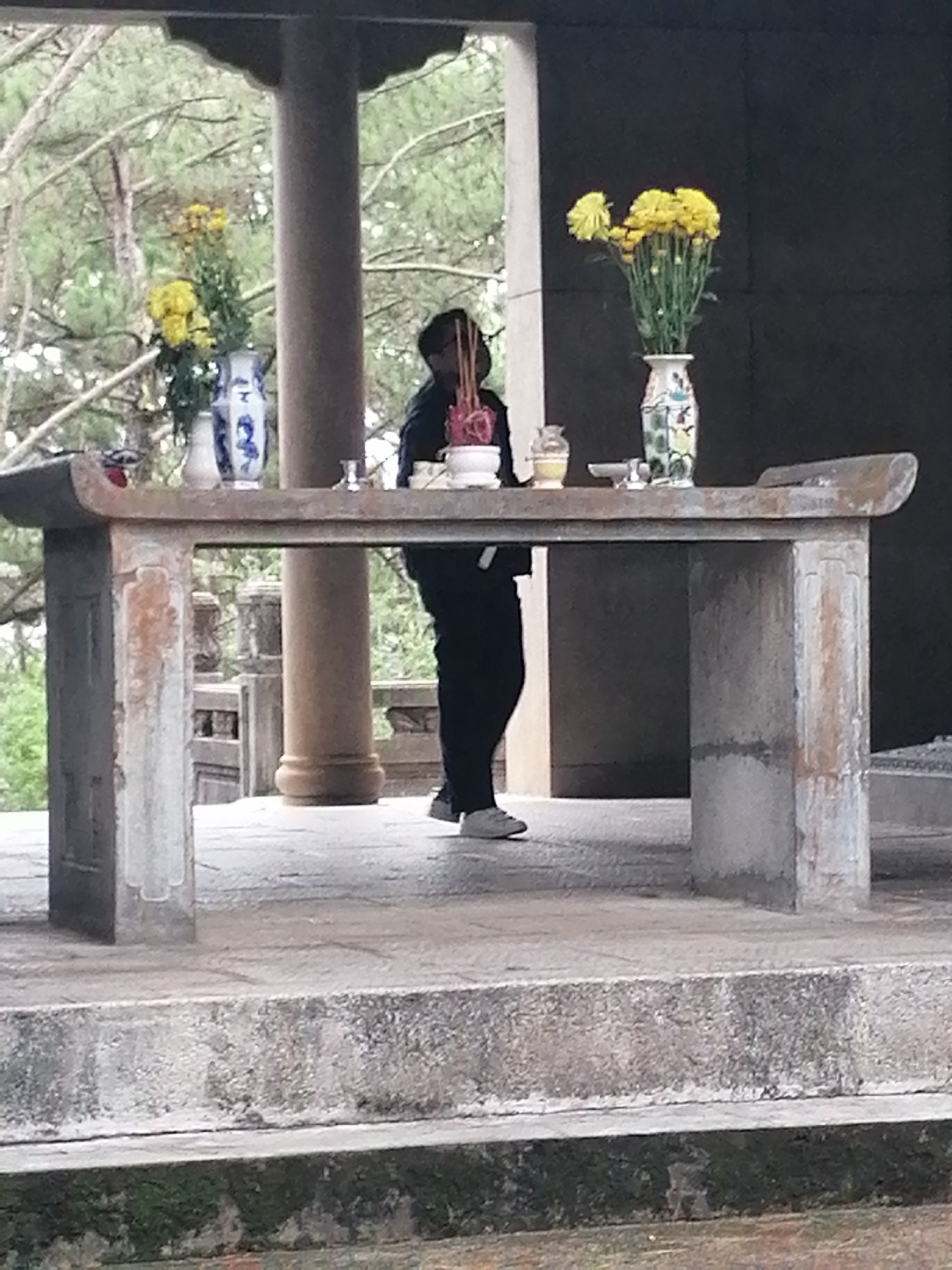 The image depicts an outdoor spiritual or ceremonial setting, likely within Cam Ly, Đà Lạt, Vietnam. A single individual stands behind a weathered, light-grey stone table or altar, engaged in what appears to be a religious or commemorative practice.
The stone table is prominent in the foreground, featuring sturdy legs and a slightly curved top surface. Arranged upon it are several offerings: two ceramic vases containing bunches of bright yellow flowers. One vase is white with blue patterns, while the other features colorful motifs. Centrally, a white bowl holds numerous red-tipped incense sticks, many of which are smoking, indicating a recent or ongoing ritual. Other small vessels are also visible on the table.
The person, silhouetted against a lighter background, is dressed in dark trousers and a dark long-sleeved top, with light-colored shoes. Their hands are held together in front of them in a gesture consistent with prayer or reverence.
The setting is an open-air structure with robust stone columns and paved flooring. Beyond the immediate architectural elements, a lush, blurred background of trees, likely pine, suggests a natural, perhaps mountainous or hilly environment. The scene is illuminated by natural daylight, conveying a calm and contemplative atmosphere. There is no visible text in the image.