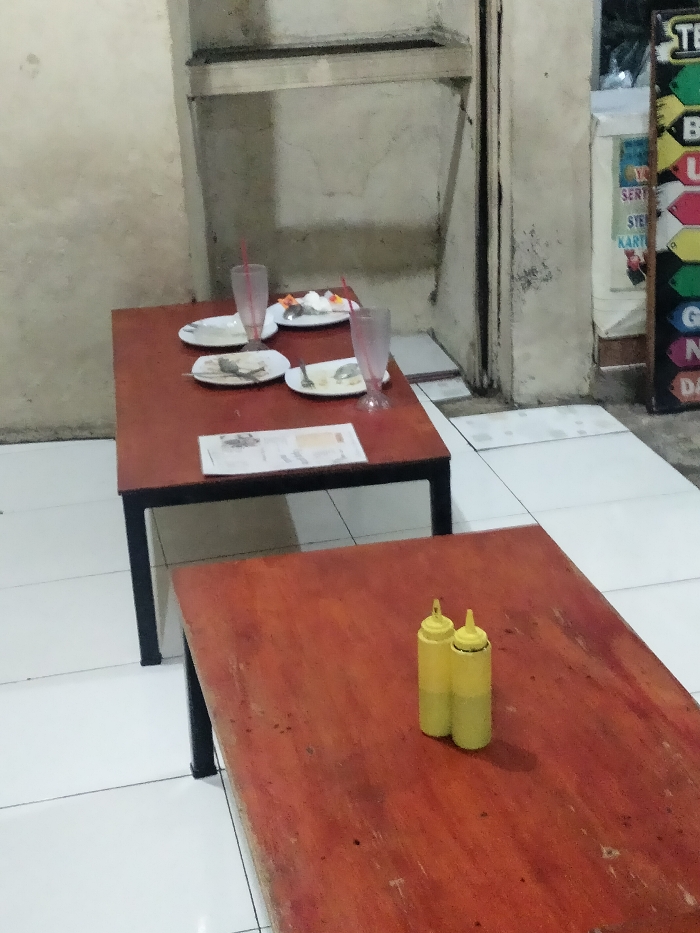 This image captures a simple, casual dining setting, likely in an eatery or warung in Dramaga, Indonesia. There are no people visible.
In the foreground, a rustic wooden table with dark metal legs features two identical yellow squeeze bottles, commonly used for condiments like chili sauce or ketchup.
In the mid-ground, another similar wooden table displays the aftermath of a meal. Four white plates, some with remnants of food and used cutlery, suggest recent dining. Two clear plastic glasses with red straws are also on the table. A white paper, possibly a menu or bill, lies flat.
The setting includes light-colored, somewhat grimy walls and a white tiled floor, indicating a well-used space. To the right, a narrow doorway or opening is partially visible next to a colorful vertical sign. Text on the sign, fragmented due to the crop, includes "SERTI", "KART", "N", and "DA", confirming an Indonesian context. The dim, artificial lighting suggests it is either evening or night. The scene portrays a functional, unpretentious dining environment after customers have departed.