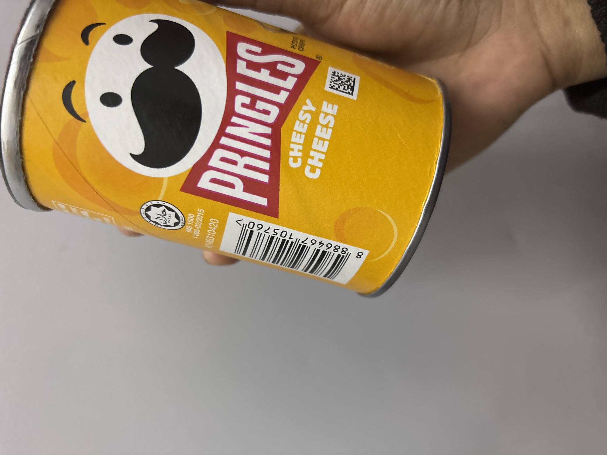 A hand holds a yellow can of Pringles Cheesy Cheese potato crisps. The Pringles logo and name, the "Cheesy Cheese" flavor designation, a halal symbol, and a barcode are visible on the can. The can's details include "MS 1500 1095-02/2015 K146310420" and the barcode reads "8 886467 105760>". The can is positioned against a grey background. The location in the city of North Jakarta, Indonesia cannot be determined from this image.