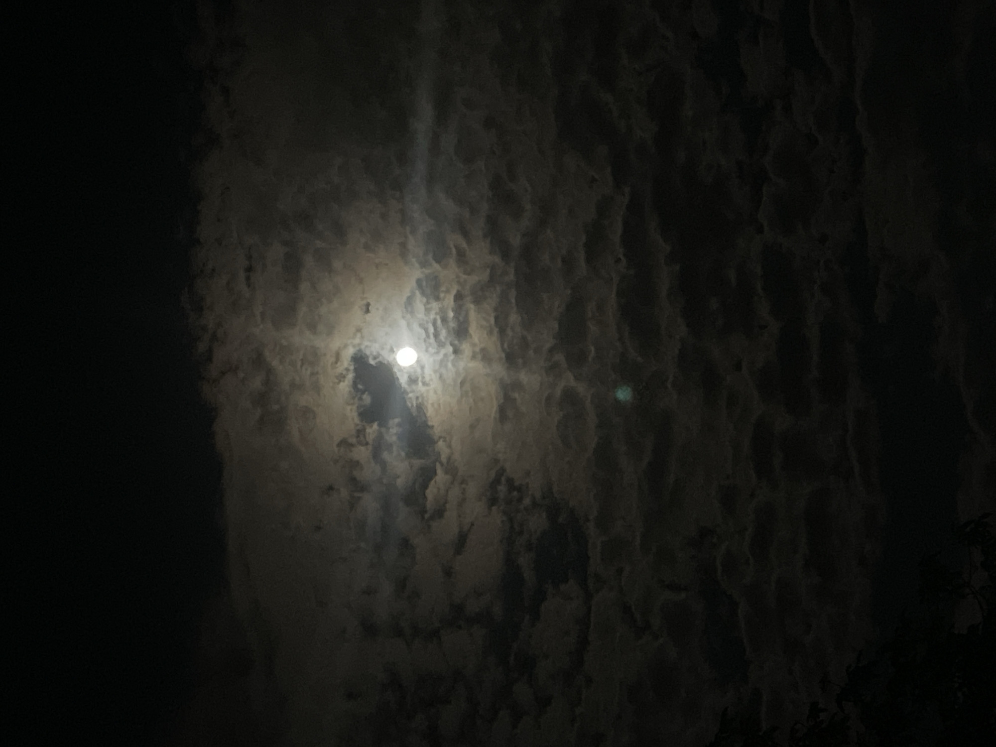 The image depicts a night sky with the moon visible through layers of dark, textured clouds. The moon is relatively small and obscured by the clouds, emitting a bright glow. A small green light is visible to the side. The image is predominantly dark. The bottom right corner of the image has tree leaves visible. The location is unknown, and there is no definitive evidence to indicate that the image was taken in Kazanlak, Bulgaria.