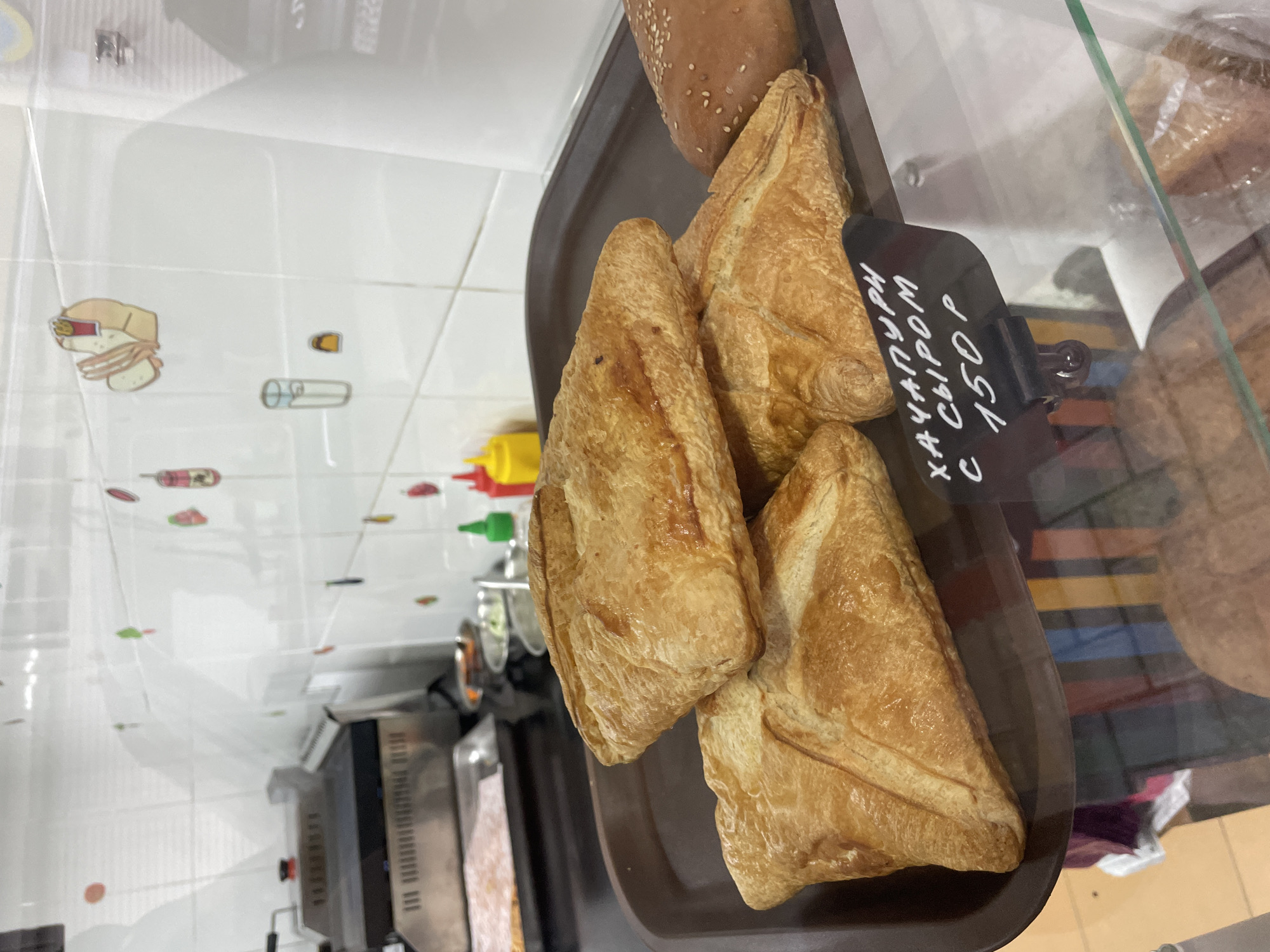 The image shows a display of baked goods, specifically triangular pastries, likely cheese-filled khachapuri, on a brown serving tray. A price tag indicates "Khachapuri with cheese, 150 Rubles." The scene appears to be within a food establishment, potentially a bakery or deli in Olginka, Russia, based on the location provided. A shelf with tiled walls with printed images, condiment bottles, and stainless steel equipment is visible in the background, suggesting a kitchen or food preparation area. The display is behind glass, indicating it is a food display case.