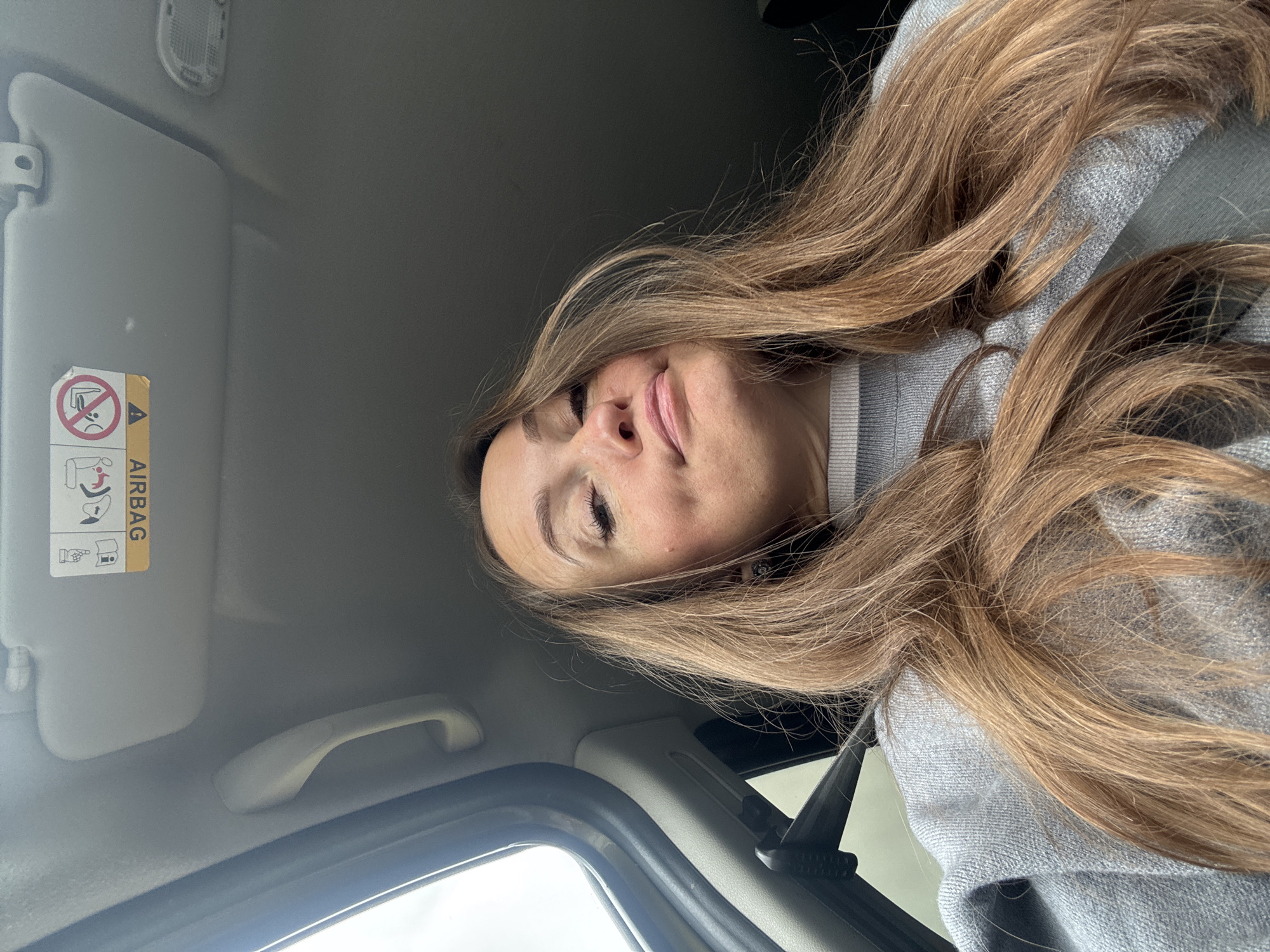 The image displays a female individual photographed from a low-angle perspective inside what appears to be a vehicle. She has long, light brown hair flowing over her shoulders and chest, and is wearing a grey, possibly knitted, sweater with a ribbed neckline. Her face is angled upwards, showing a slight smirk or pout, with her right eye appearing partially closed or squinting. Visible freckles are present on her nose and cheeks. A black seatbelt strap is seen across her left shoulder. The vehicle's interior features a light grey roof lining, a matching grab handle on the passenger side, and a sun visor. An airbag warning label is affixed to the sun visor, with the text "AIRBAG" appearing both horizontally mirrored and upside down in relation to the image frame. A dome light or similar overhead console fixture is partially visible in the upper right corner. Through the driver's side window, an overcast sky is discernible. The specific location, including the city of Поварёнки, Russia, cannot be determined from the visual content of the image.