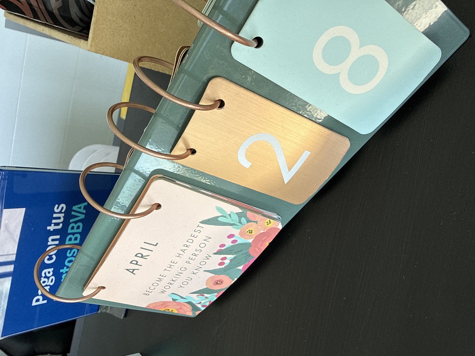 The image displays a desktop perpetual calendar set to April 28, tilted on a dark surface, likely a desk. The calendar features a ring binding made of a copper-toned material, holding three distinct cards. The uppermost card is light blue with a large white number '8', partially obscured. Below it, a metallic gold or bronze card displays a large white number '2'. The bottom card, made of light pink or beige material, shows the word "APRIL" at the top. Underneath, a motivational quote reads: "BECOME THE HARDEST WORKING PERSON YOU KNOW". This card is adorned with a floral illustration featuring pink, orange, and green hues along its bottom edge.
To the left of the calendar, a blue object, possibly a brochure or folder, is partially visible. Text on this object, in Spanish, reads "Paga con tus tos BBVA" and "Hando Oportunidades", suggesting a connection to banking services, specifically BBVA. A brown, cardboard-like box is visible in the upper left corner, and a plain light-colored wall or surface is in the background. The overall scene suggests an indoor setting, consistent with an office or home environment in San Bernardino Tlaxcalancingo, Mexico, as indicated by the Spanish text from BBVA.