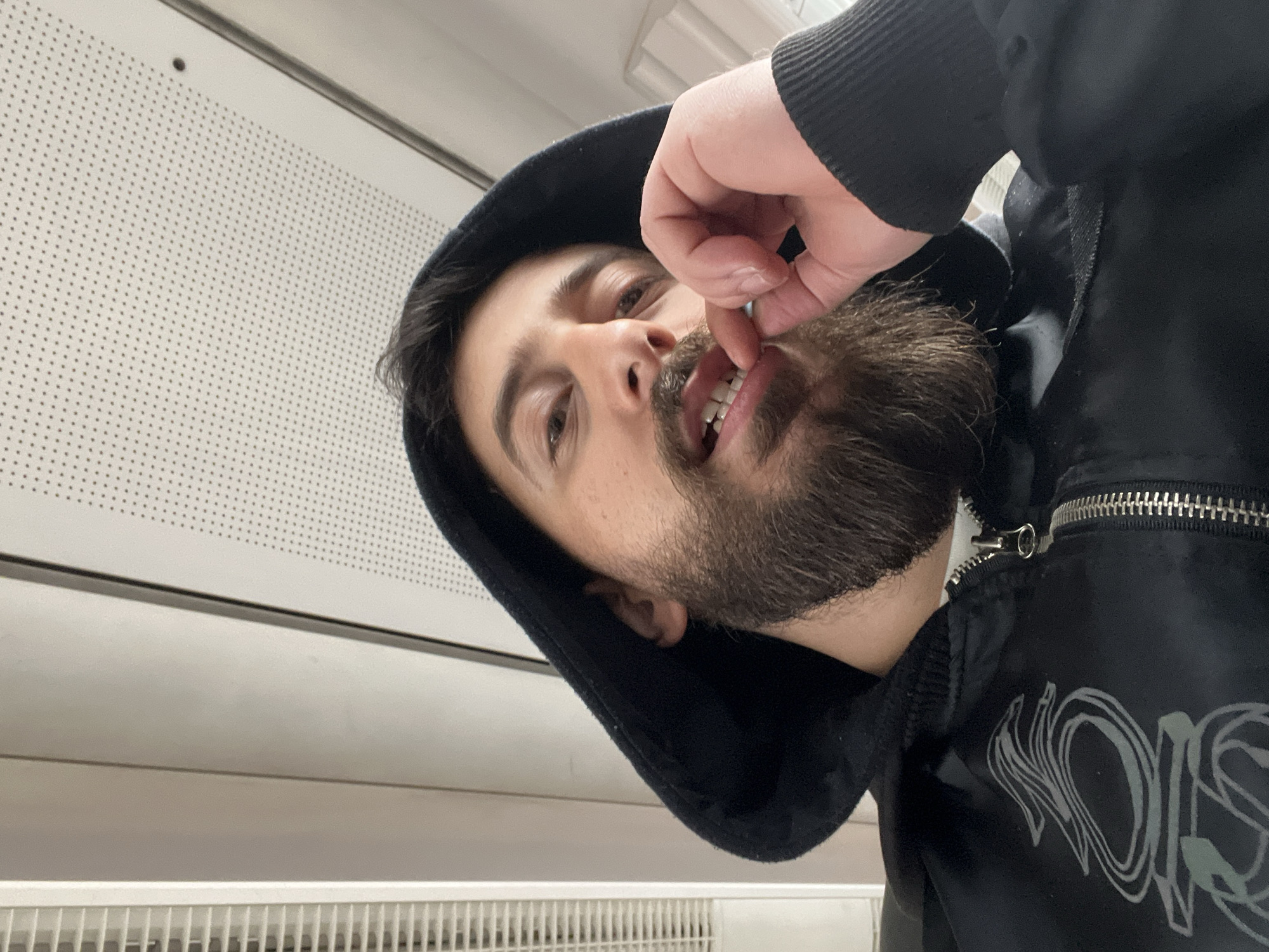 A close-up, tilted, self-portrait photograph shows a man wearing a black hooded jacket. He has dark hair and a thick beard, and he is picking at his teeth with his fingers. The background consists of white perforated paneling above a radiator. The location is unknown; there is no indication of Strasbourg, France.