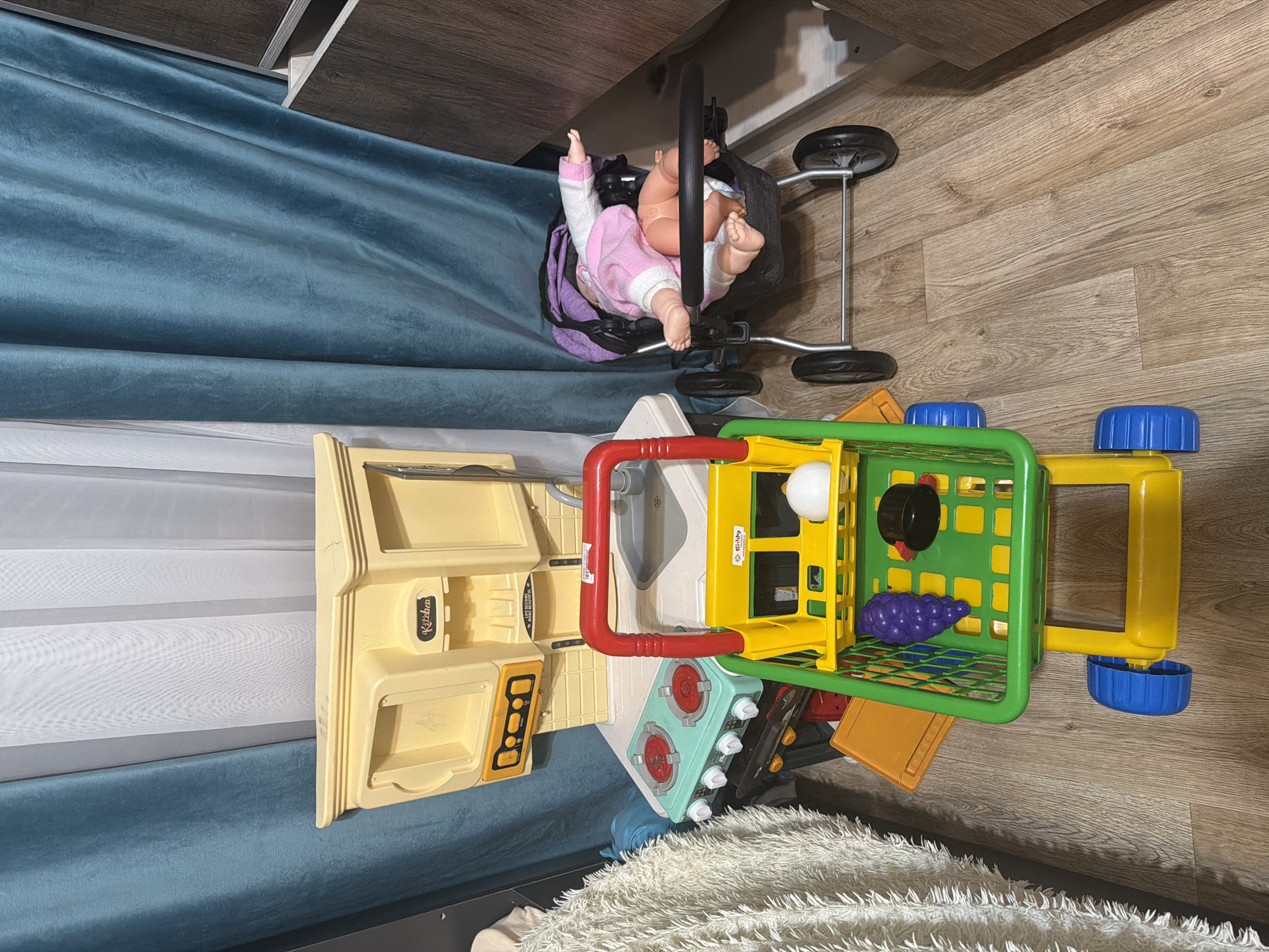 The image shows a collection of children's toys arranged on a wooden floor. In the foreground, a yellow and green toy shopping cart is filled with various toy food items, including purple grapes and a white spherical object, possibly an egg or onion. A black cylindrical object, perhaps a pot or container, is also present.
Behind the shopping cart, a toy kitchen set is visible, predominantly yellow, with red elements and markings that suggest buttons or controls. To the right of the kitchen set, a toy baby doll is lying in a black baby stroller with a purple and pink blanket. The doll is wearing a pink and white outfit.
In the background, blue and white curtains are draped. A fluffy white textured object, likely a rug or blanket, is partially visible on the left side of the image. The overall environment appears to be an indoor setting, possibly a child's playroom or bedroom. There are no visible signs or indicators that would suggest the specific location in the city of Shymkent, Kazakhstan.