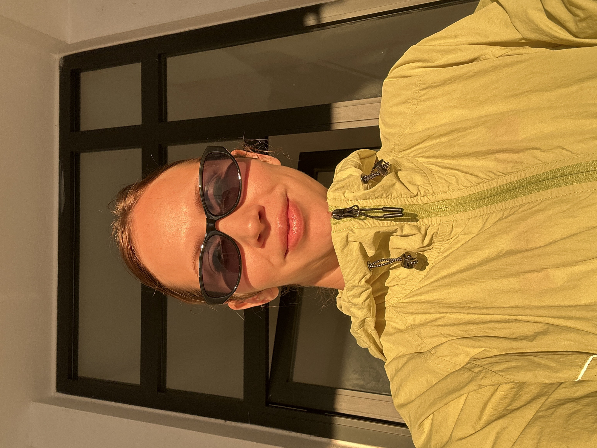 A woman wearing sunglasses and a light yellow jacket poses in front of a window with black frames. The light suggests it is either early morning or late afternoon. The location cannot be determined to be Fethiye, Turkey, based on the information in the image.