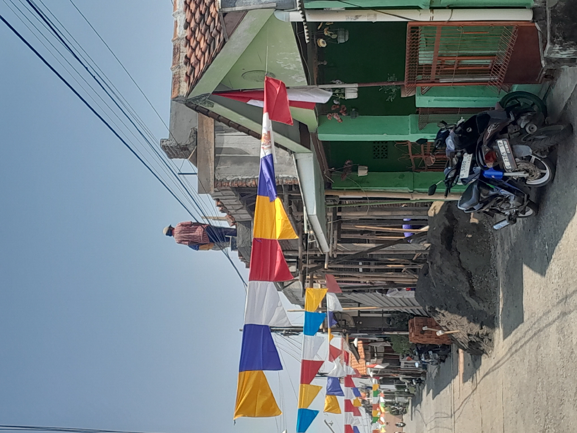 A street in Kalijeruk, Indonesia is lined with buildings and decorated with colorful flags. Two men are on the roof of a building, one in a striped shirt and the other in a blue shirt. There are several motorcycles parked on the side of the road. A pile of dirt and bricks is in the foreground. The sky is clear and blue.