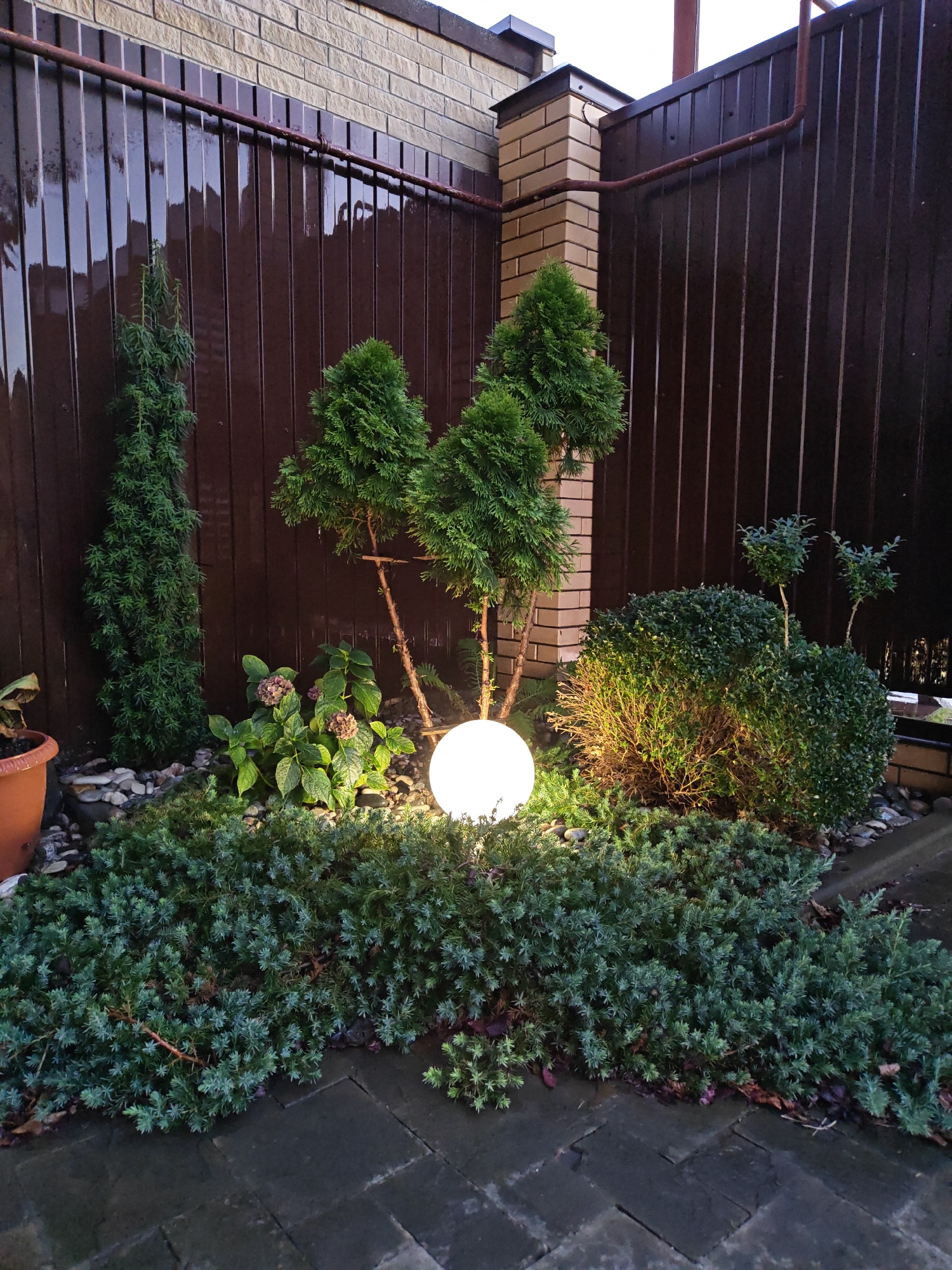 This image shows a small, well-maintained garden area in Vodopadnyy, Russia, adjacent to a tall, dark brown corrugated metal fence and a light brick pillar. The garden features several types of evergreen shrubs and trees, including a tall, slender evergreen on the left, three conical evergreens in the center, and a large, rounded shrub on the right. A bright, spherical light fixture is positioned in the center of the garden, illuminating the surrounding plants and ground. The ground is covered with dark grey interlocking paving stones and decorative pebbles. A terracotta pot with a dead plant is visible on the far left. The overall scene suggests a private backyard or garden space, likely illuminated for evening ambiance.