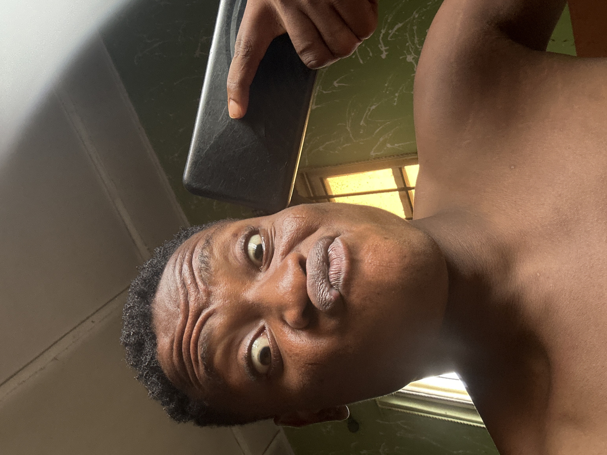 A person is taking a selfie with a smartphone in a room. The person's face is close to the camera, with eyes wide and lips pursed. The person is shirtless, and a portion of their shoulder and neck are visible. The background shows a green wall with a white pattern and a window with yellow light streaming through. A portion of the smartphone, held by a hand with an extended finger, is visible in the upper right corner of the image. The scene is likely an indoor residential setting in Ikorodu, Nigeria.
