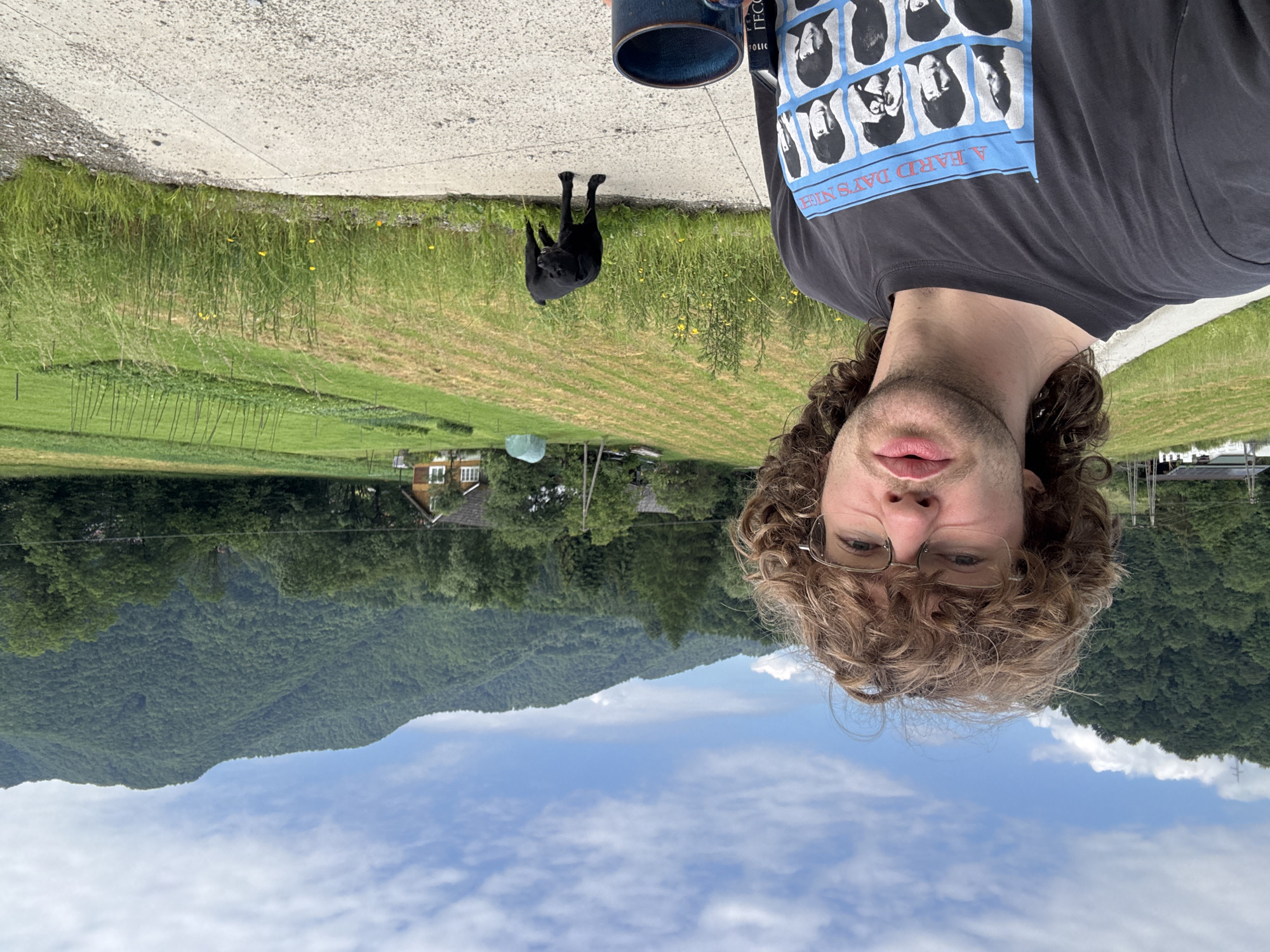 A young man with curly brown hair and glasses is taking a selfie outdoors. He is wearing a dark gray t-shirt with a graphic of the Beatles from "A Hard Day's Night." In his left hand, he holds a blue mug. To his right, a black dog stands on a concrete path. Behind them, a field with rows of crops stretches towards a cluster of buildings, including a house with a red roof and white trim, and a greenhouse. Rolling green hills and a dense forest are visible in the background under a cloudy blue sky. The location is described as Unknown, Unknown.