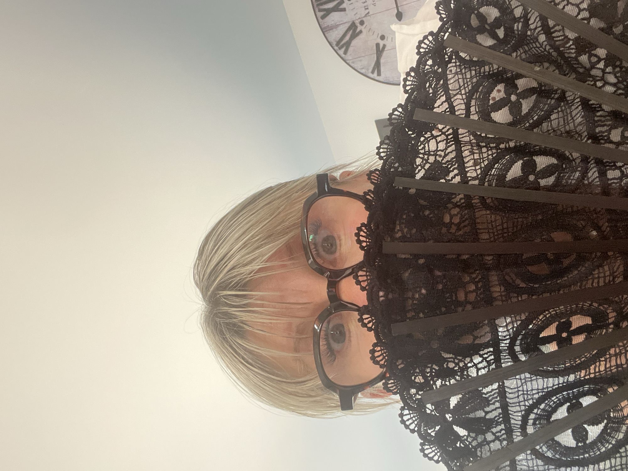 This is a portrait of a woman in Chambœuf, France. The image is oriented vertically with the top of the image on the left. The woman has short, blonde hair and is wearing glasses with black frames. She is holding a black lace fan in front of her face, obscuring part of it. Her eyes are visible through the fan. Behind her, a clock with Roman numerals can be seen on the wall, indicating the time is approximately 3:40. The background appears to be an indoor setting with light-colored walls. The overall lighting suggests it is daytime. The woman's expression is neutral. There is no visible text in the image apart from the numerals on the clock.