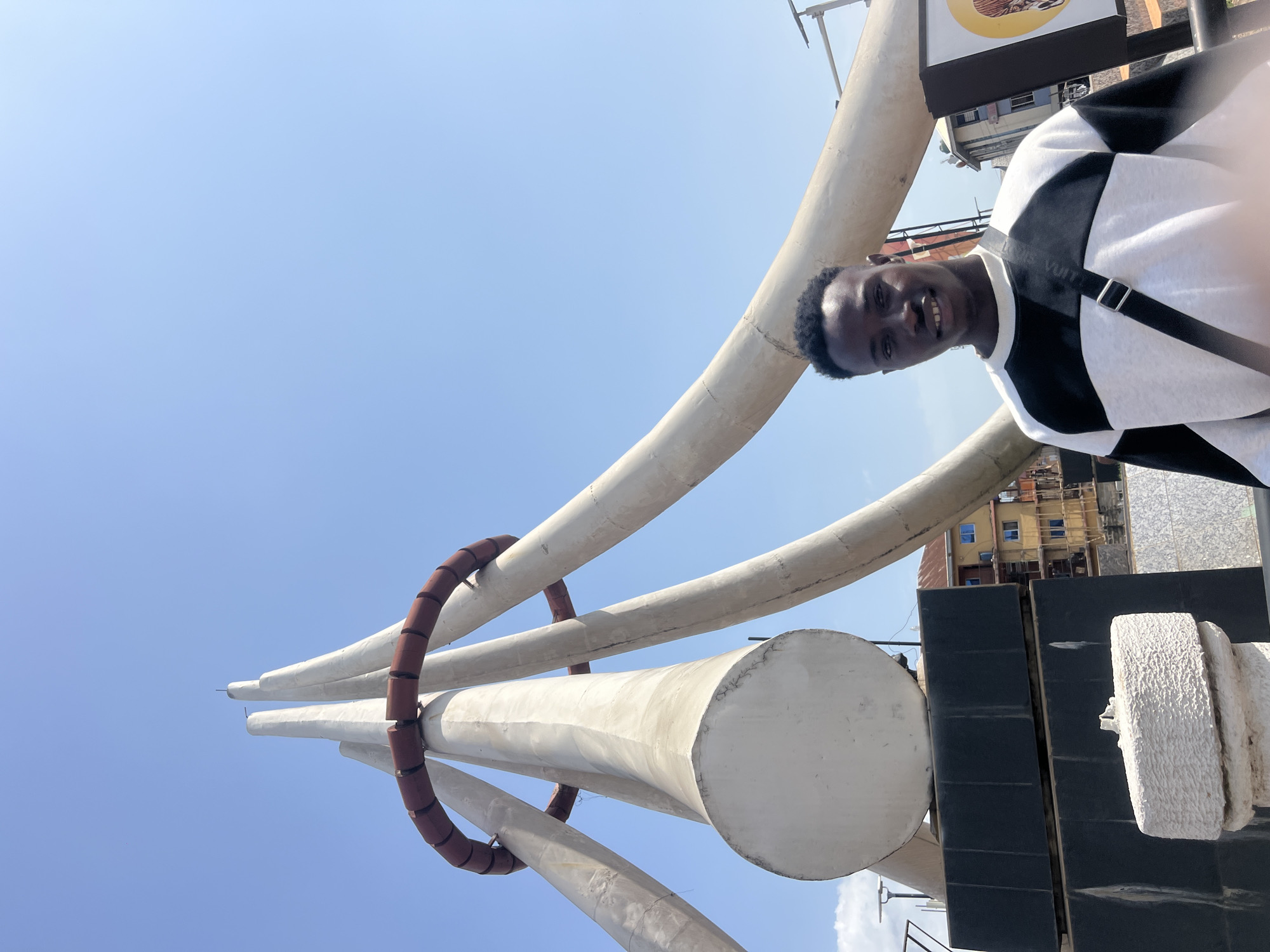 A young man with dark skin and a broad smile poses for a photo, likely a selfie, in front of a monumental sculpture under a clear blue sky. He wears a black and white long-sleeved top with a diagonal color block pattern. A black strap, faintly bearing "LOUIS VUITTON" in white text, crosses his right shoulder.
The dominant feature of the scene is a large sculpture composed of multiple light-colored, possibly concrete or stone, horn-like or tusk-like structures that curve upwards and converge. A prominent red, segmented ring encircles several of these structures mid-way, and another smaller red ring is visible higher up. The base of the sculpture is dark, appearing to be made of rectangular blocks, with some rough-textured white elements at its foot.
In the background, behind the sculpture, parts of urban architecture are visible. This includes multi-story buildings, some with blue-tinted windows, suggesting a commercial or residential area. A commercial sign with a yellow/orange background and partial black text (an 'O' is discernible) is also visible. The bright lighting indicates it is daytime, under sunny weather. The setting is an outdoor public space, identified as Onitsha, Nigeria. The man's joyful expression conveys happiness and comfort.