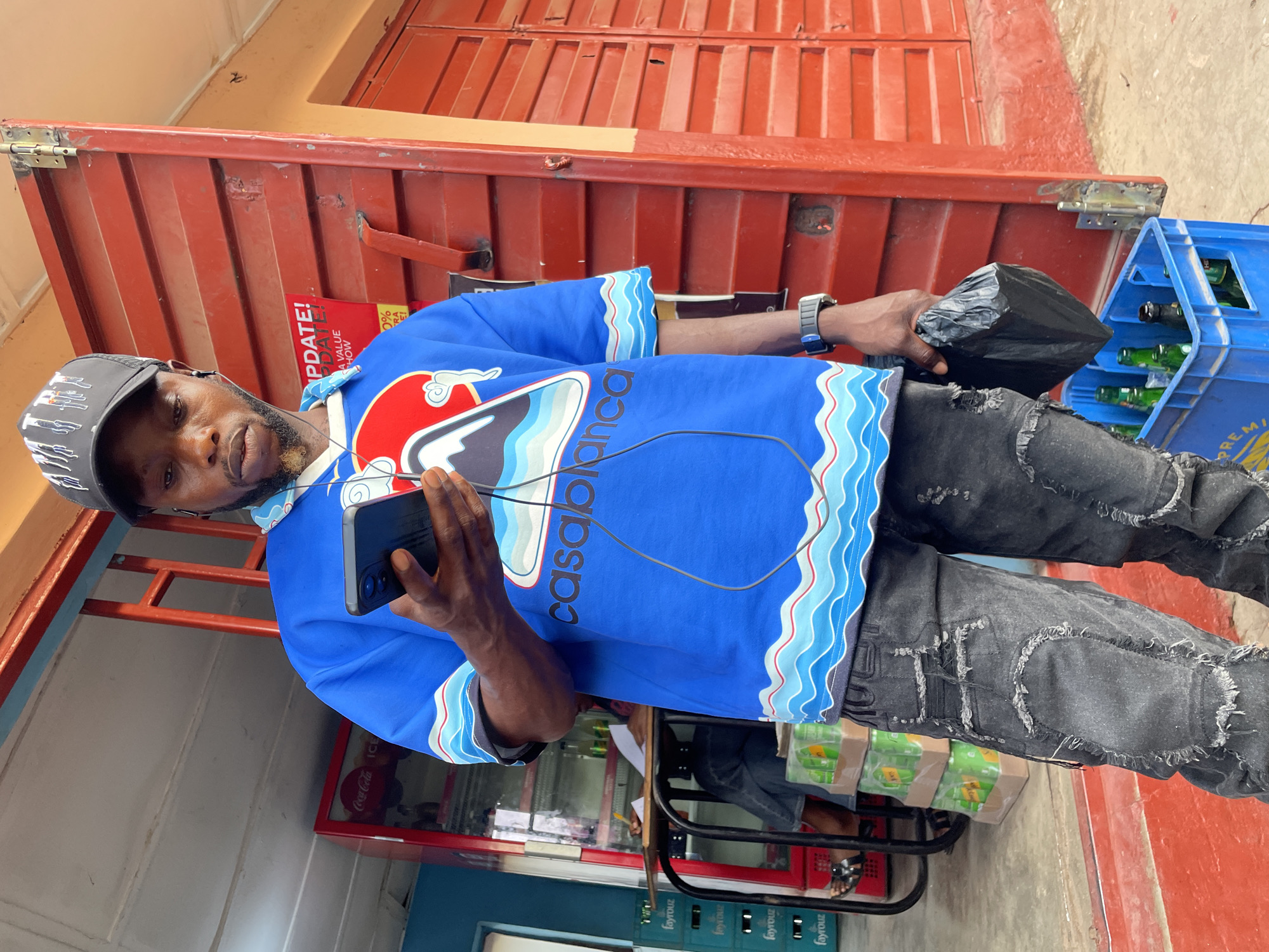 An outdoor, slightly rotated, medium shot captures a man standing in what appears to be a street or market setting in Jos, Nigeria. He is centered in the frame, wearing a blue short-sleeved shirt with "casablanca" and wave patterns, ripped black jeans, and a black baseball cap. Earphones are visible around his neck and connected to a black smartphone he holds in his left hand, while his right hand holds a black plastic bag. A black wristwatch is on his right wrist.
The background features a red, corrugated metal structure, possibly a kiosk or container, with hinges and latches visible. To the left, a red Coca-Cola branded fridge is partially visible. To the right, two blue plastic crates filled with green bottles are stacked. Green cardboard boxes, some labeled "fayrouz," are stacked on a metal stand below the crates.
Visible text includes "Coca-Cola ICE" on the fridge, "UPDATE!", "PDATE!", "A VALUE", "HOW", "0%", "RA" on a red sign attached to the metal structure, "casablanca" on the man's shirt, and "PREMI" on one of the blue crates. The man has a neutral expression, looking directly at the camera. The scene is brightly lit, suggesting daytime.