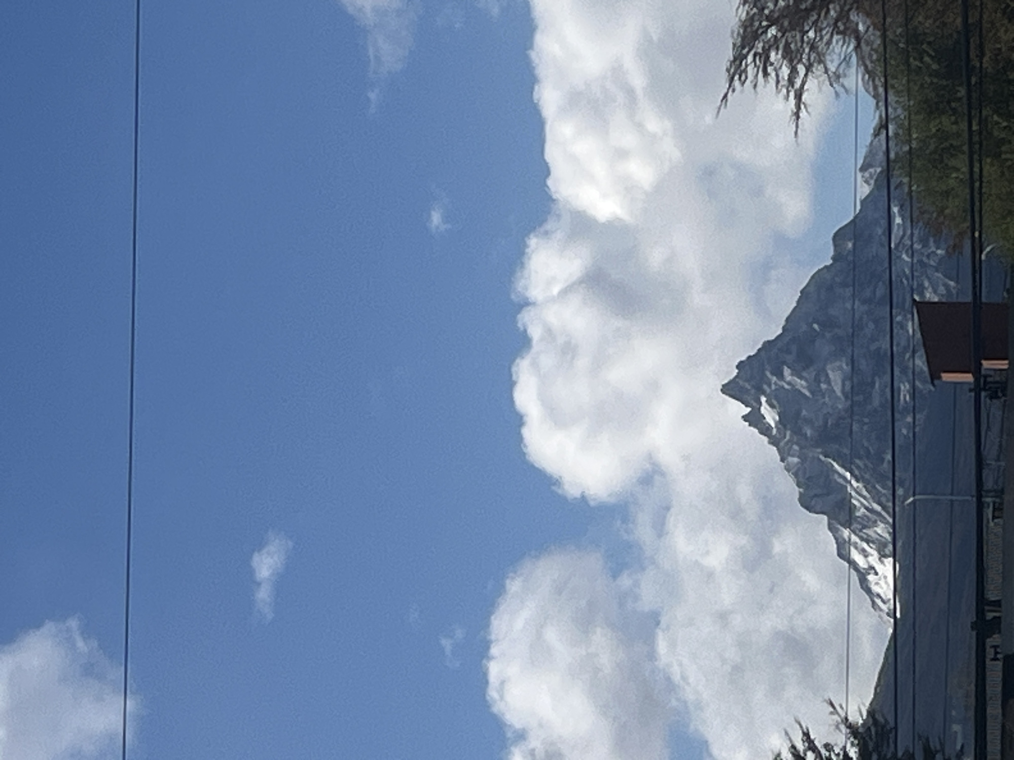 The image shows a partial view of the sky over Ushuaia, Argentina, with a prominent mountain range partially obscured by clouds on the right side of the frame. Clear blue sky dominates the left side, punctuated by a few scattered clouds. Power lines are visible vertically along the left edge.