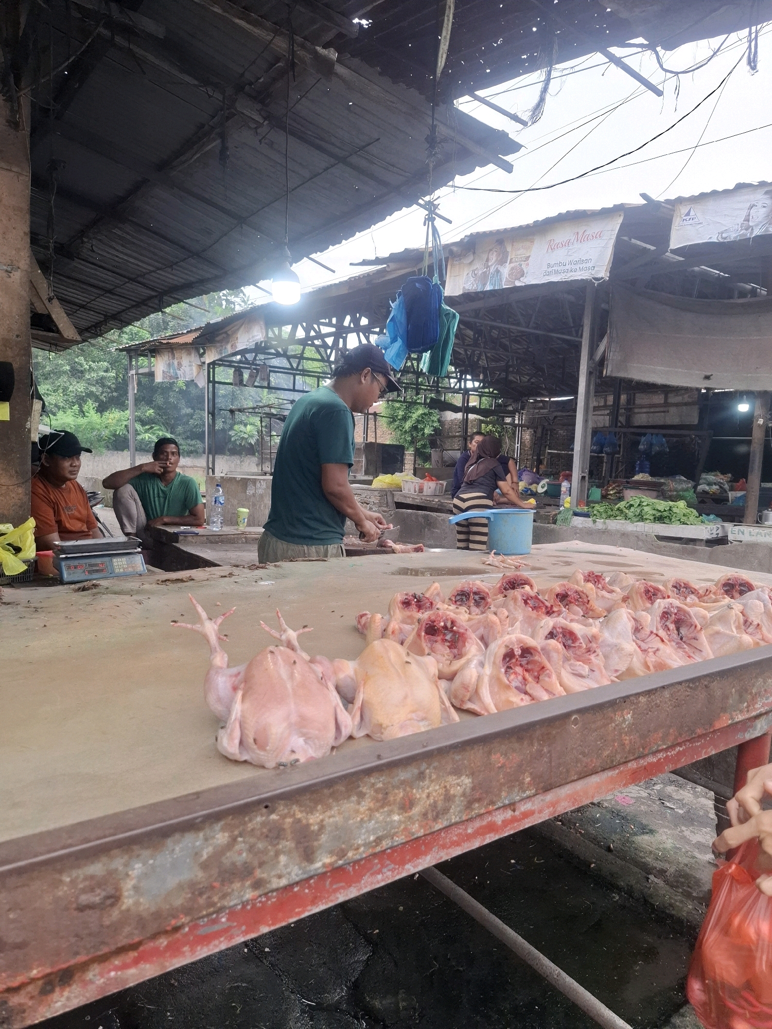 At a street-side market in Kenangan, Indonesia, a butcher, wearing a green t-shirt and a blue cap, is preparing raw chickens laid out on a large, worn metal table. The chickens are arranged in rows, some whole and others prepared for sale. Flies are visible on some of the chickens. Behind the butcher, two men are seated; one in an orange t-shirt, the other in a green t-shirt. A digital scale is on the table in front of the man in the orange shirt. The market appears to be under a makeshift roof supported by wooden poles, with electrical wires and hanging bags visible. In the background, other market stalls are set up, with produce and more people engaged in market activities. The overall environment suggests an open-air market with functional but rustic infrastructure.