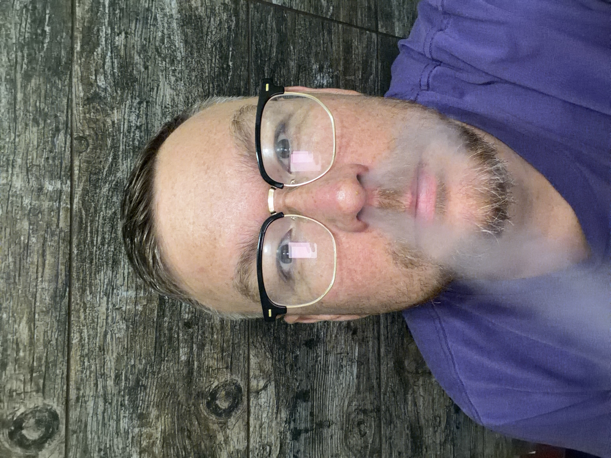 The image is a close-up, head-and-shoulders shot of a man wearing glasses and a purple t-shirt. He has light brown hair, fair skin, and a light beard. He appears to be exhaling smoke, which obscures his mouth and chin. The background is a wooden wall with a visible grain pattern. The image does not contain any information that would allow for the identification of a specific location in Kazan, Russia.