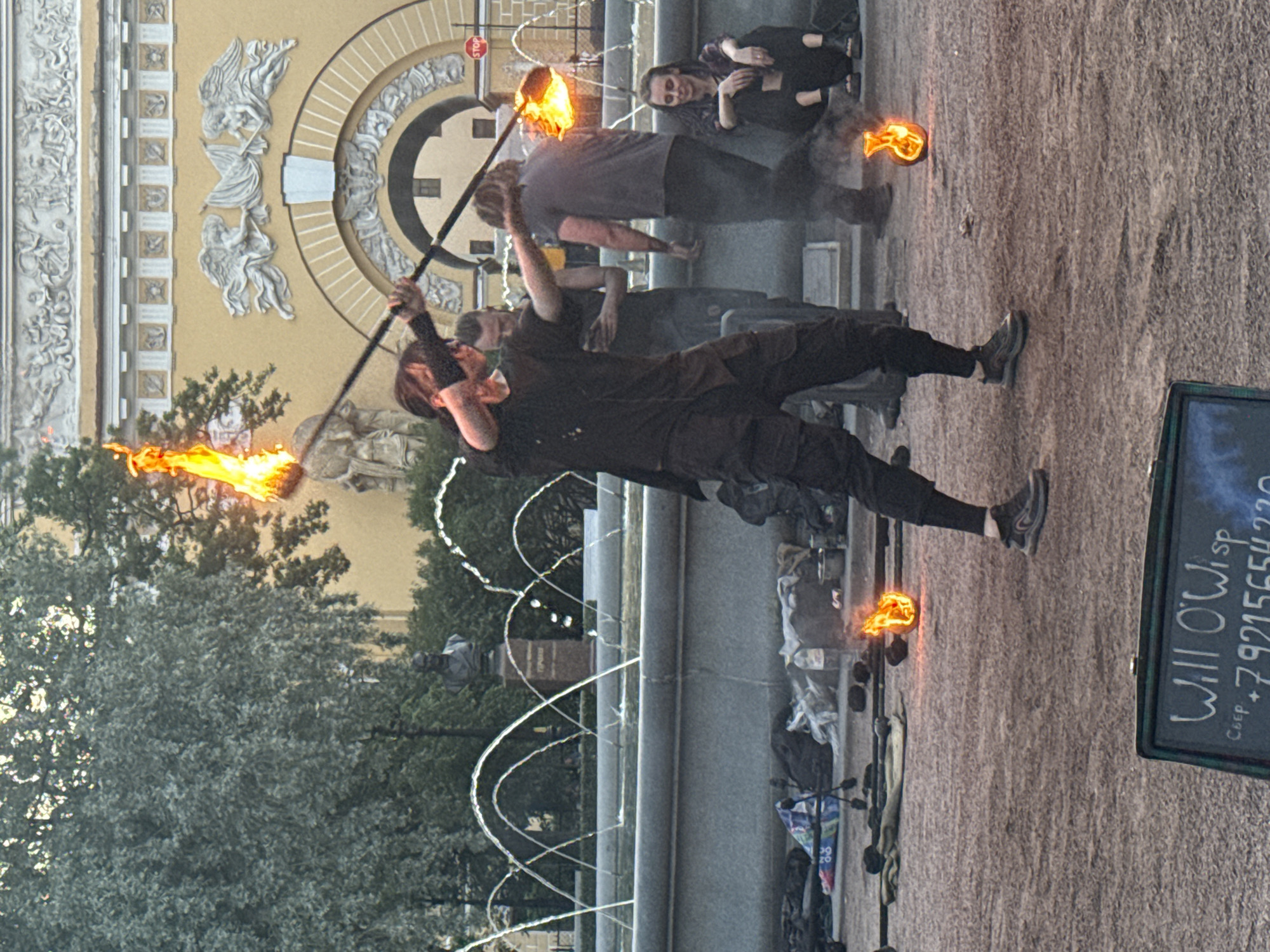 The image depicts a street performance involving fire dancing in front of the General Staff Building in Palace Square, Saint Petersburg, Russia. The central figure is a fire performer in dark clothing, actively twirling a flaming staff. Several other individuals are present, some observing the performance and others seemingly involved in the setup or audience. A fountain is visible in the background, with water jets arching through the air, and the ornate facade of the General Staff Building serves as the backdrop. In the foreground, a sign with the text "Will O'Wisp" and a phone number is visible, suggesting a performance or service being advertised. Scattered on the ground are various props and belongings, including bags, bottles, and fire-related equipment. The ground surface appears to be a mixture of dirt and gravel. The overall scene captures a lively outdoor event with elements of artistic performance and urban landscape.