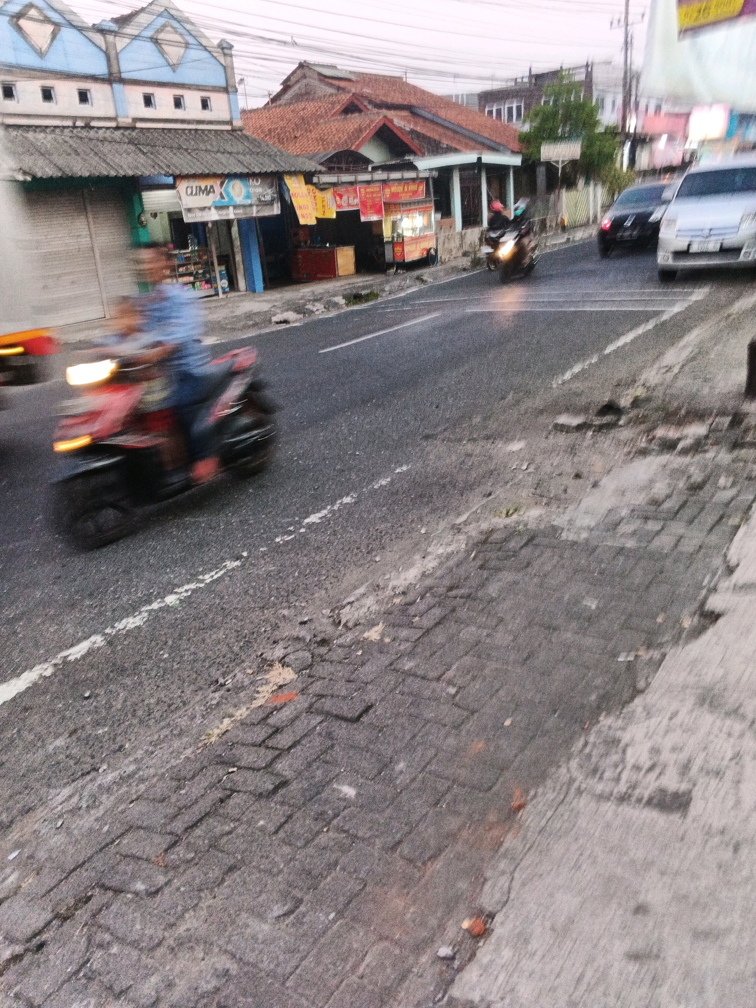 A blurred, eye-level, medium shot captures a street scene in Ungaran, Indonesia, at dusk. A person on a red and black scooter with its headlights illuminated is in motion, creating a motion blur. The scooter has an orange seat and black wheels. To the left, a building with a light blue facade and pointed roof elements is visible, featuring several small, dark windows and a gray corrugated roof. A closed metal shutter spans the lower part of this building. Further to the right, a building with a reddish-brown tiled roof and a lower gray roofline is present. A banner with the word "CLIMA" and other unreadable text is attached to the front of this building. The street surface is asphalt with visible patches of cobblestone and some debris. In the background, other vehicles, including a black car and a silver car, are also blurred by motion. The sky above is overcast.